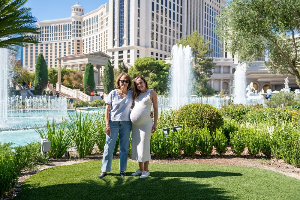 Two women stand arm in arm on a manicured lawn near sparkling fountains, with lush landscaping and a grand hotel in the background.