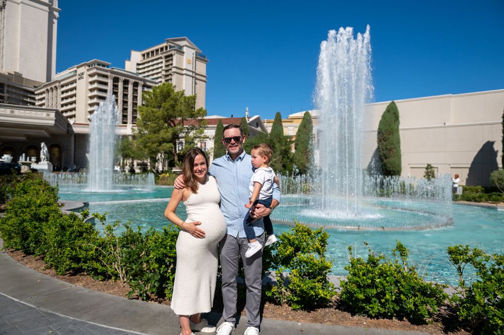 Smiling pregnant woman, man and toddler pose together in front of a large sparkling fountain with palm trees and buildings on a sunny day.