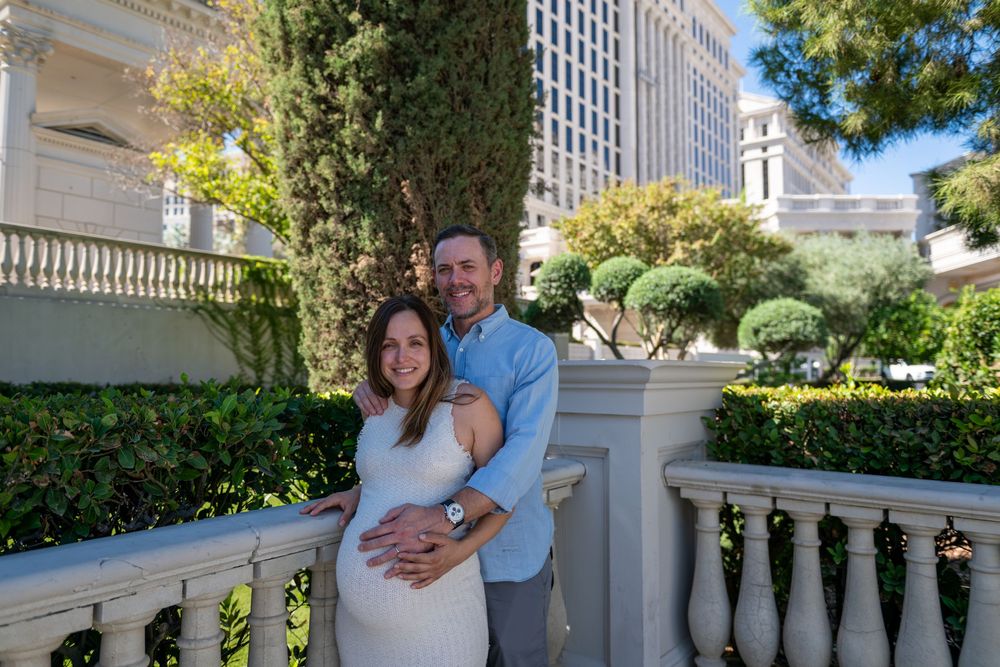 Smiling couple embraces on a white stone balcony with green hedges and tall trees, the woman in a white dress cradling her baby bump.