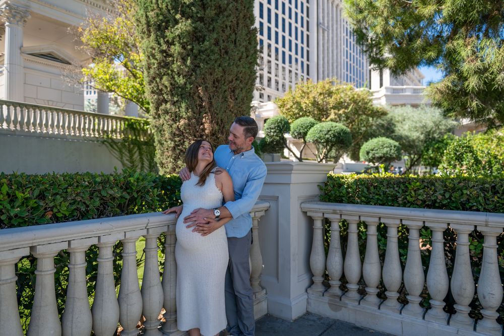 Expectant couple embracing on a stone balcony with balustrade, surrounded by manicured hedges and trees, in a sunny maternity photoshoot.