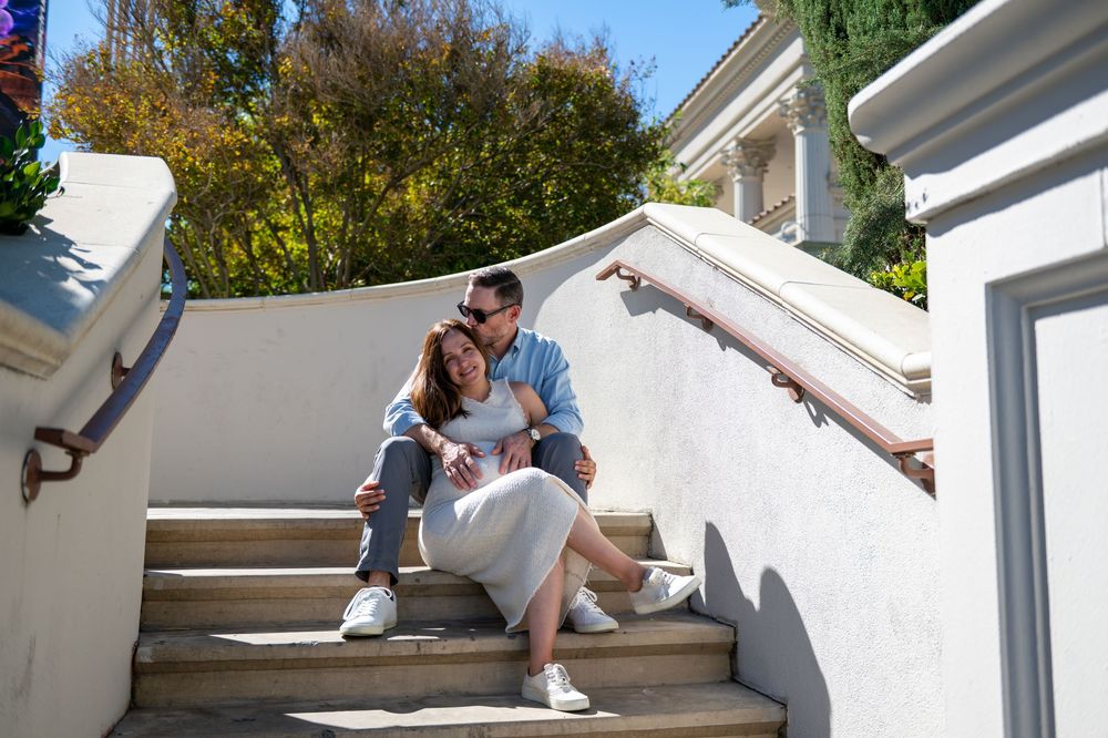Couple embracing and smiling while seated on outdoor stone steps with a railing, surrounded by trees on a sunny day.