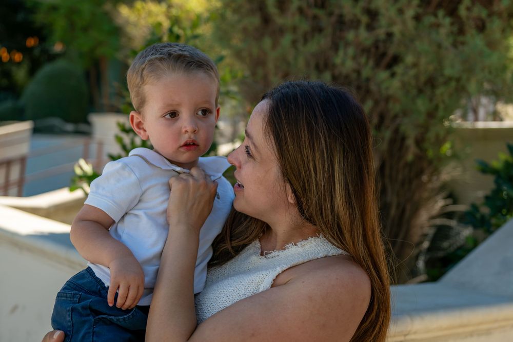 A woman with long brown hair holds a young toddler in a white shirt while standing outdoors near a stone wall and green trees.