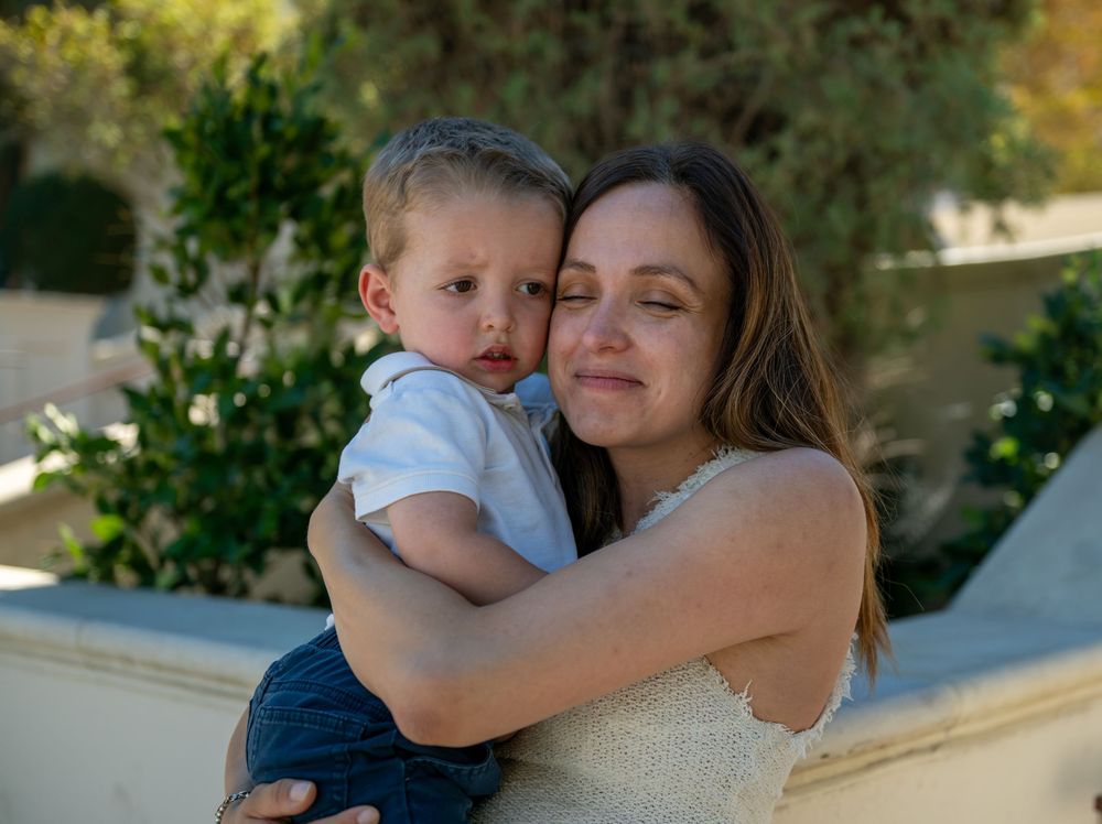 A woman with closed eyes tenderly holds a toddler in a white shirt while standing outdoors near green shrubs.