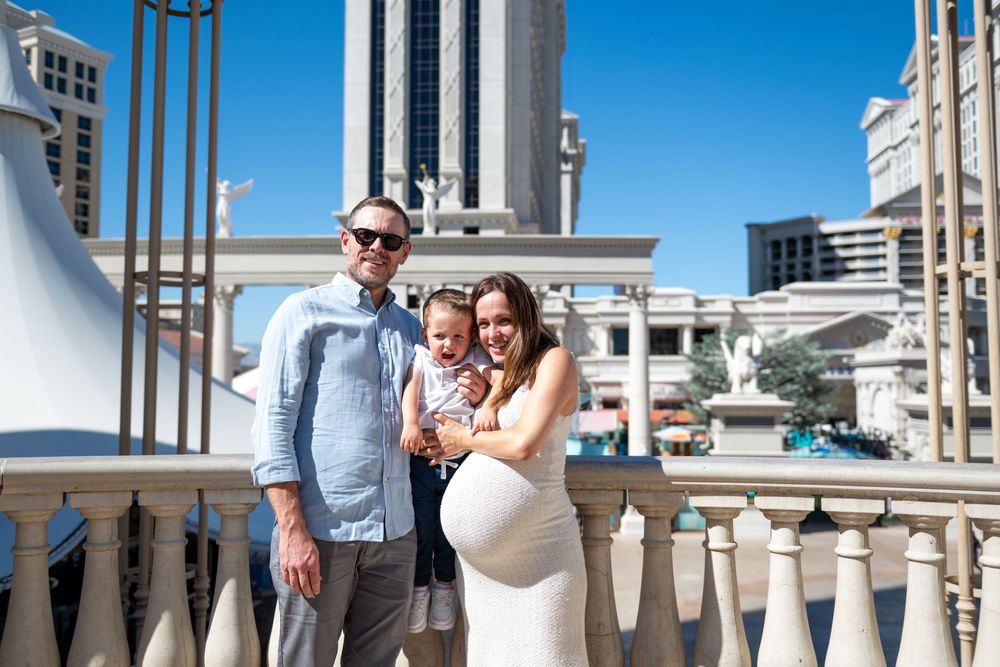 Smiling couple with a toddler posing on a stone balustrade, the pregnant woman cradling her baby bump, with Las Vegas buildings behind.