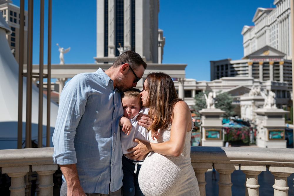 Man and pregnant woman lovingly hold a toddler between them on a Las Vegas balcony, with classical statues and tall buildings behind.