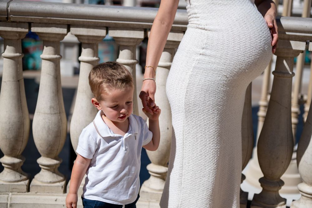 A toddler in a white polo shirt holds the hand of a pregnant woman in a fitted white dress beside a stone balustrade.