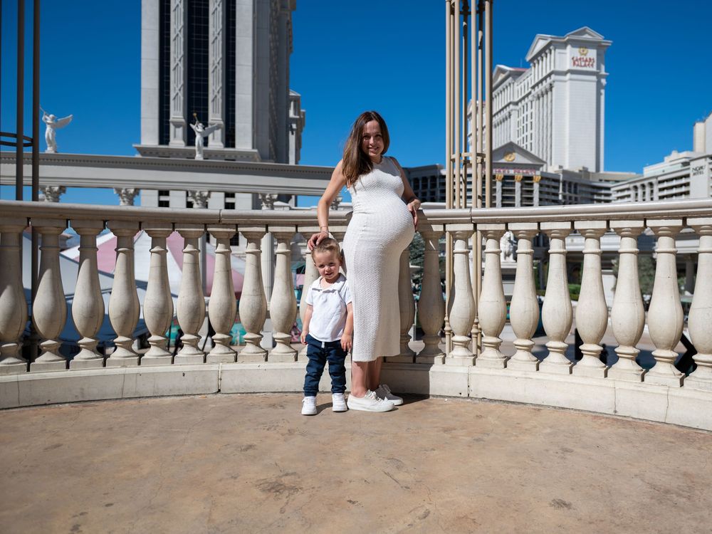 A pregnant woman in a white dress stands with her young son on a sunny stone balcony with city buildings behind.