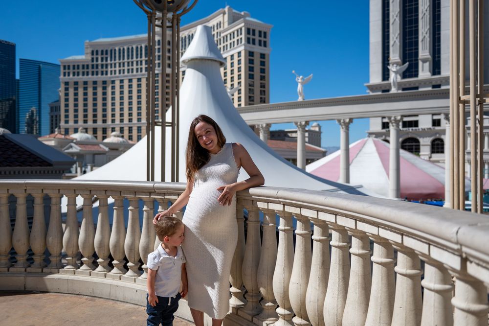 Pregnant woman in a white dress stands with a young boy on a balcony with a balustrade overlooking a sunny city skyline.