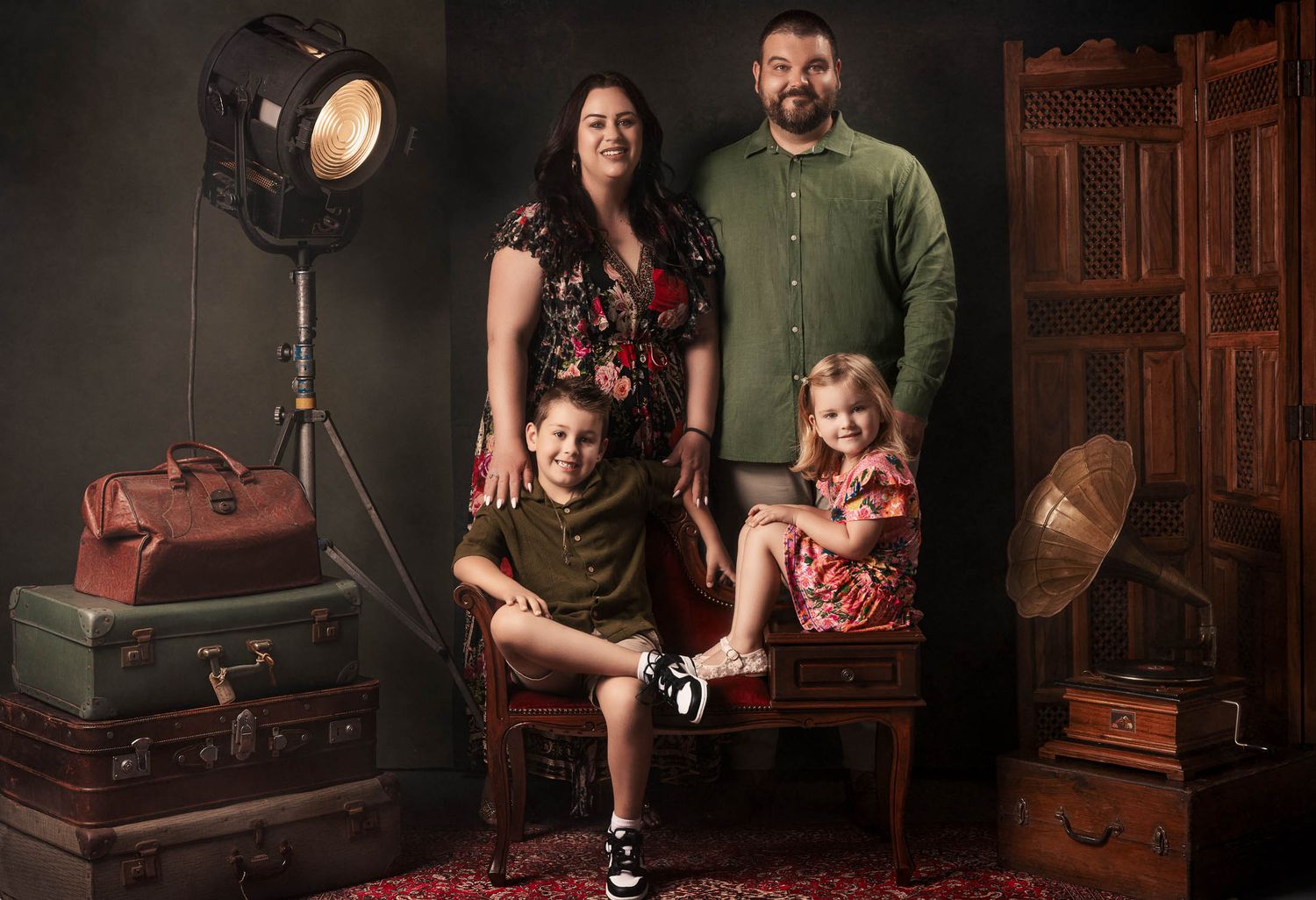 Smiling family of four poses in a vintage studio with antique suitcases, a studio light, a gramophone and a carved wooden screen.