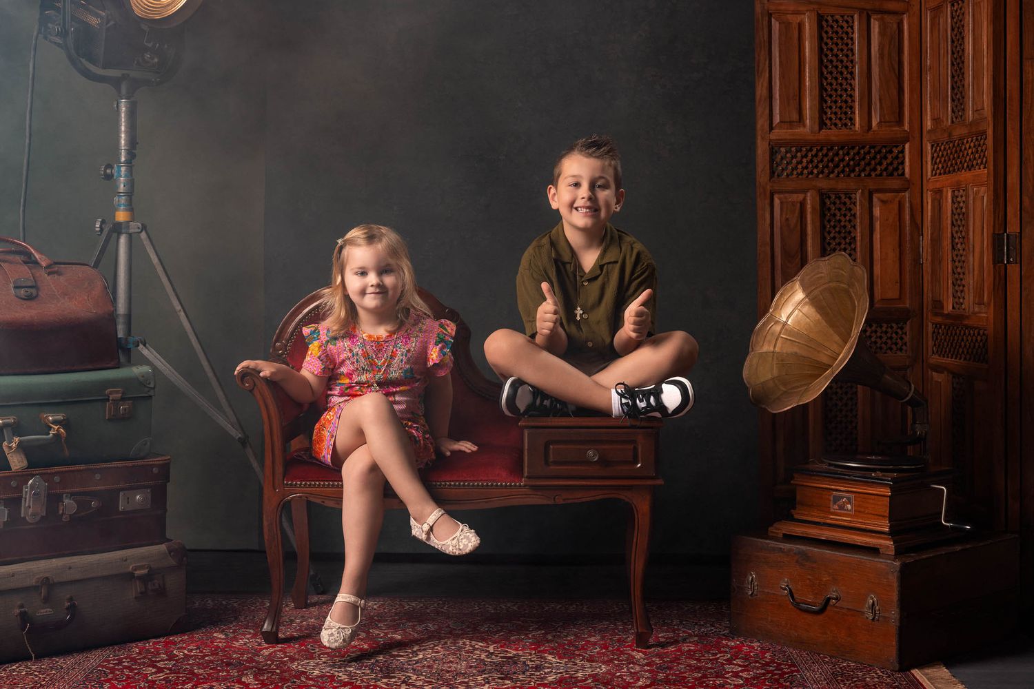 Two smiling children sit on a vintage red bench beside a brass gramophone, antique trunks and a carved wooden cabinet in a retro studio.