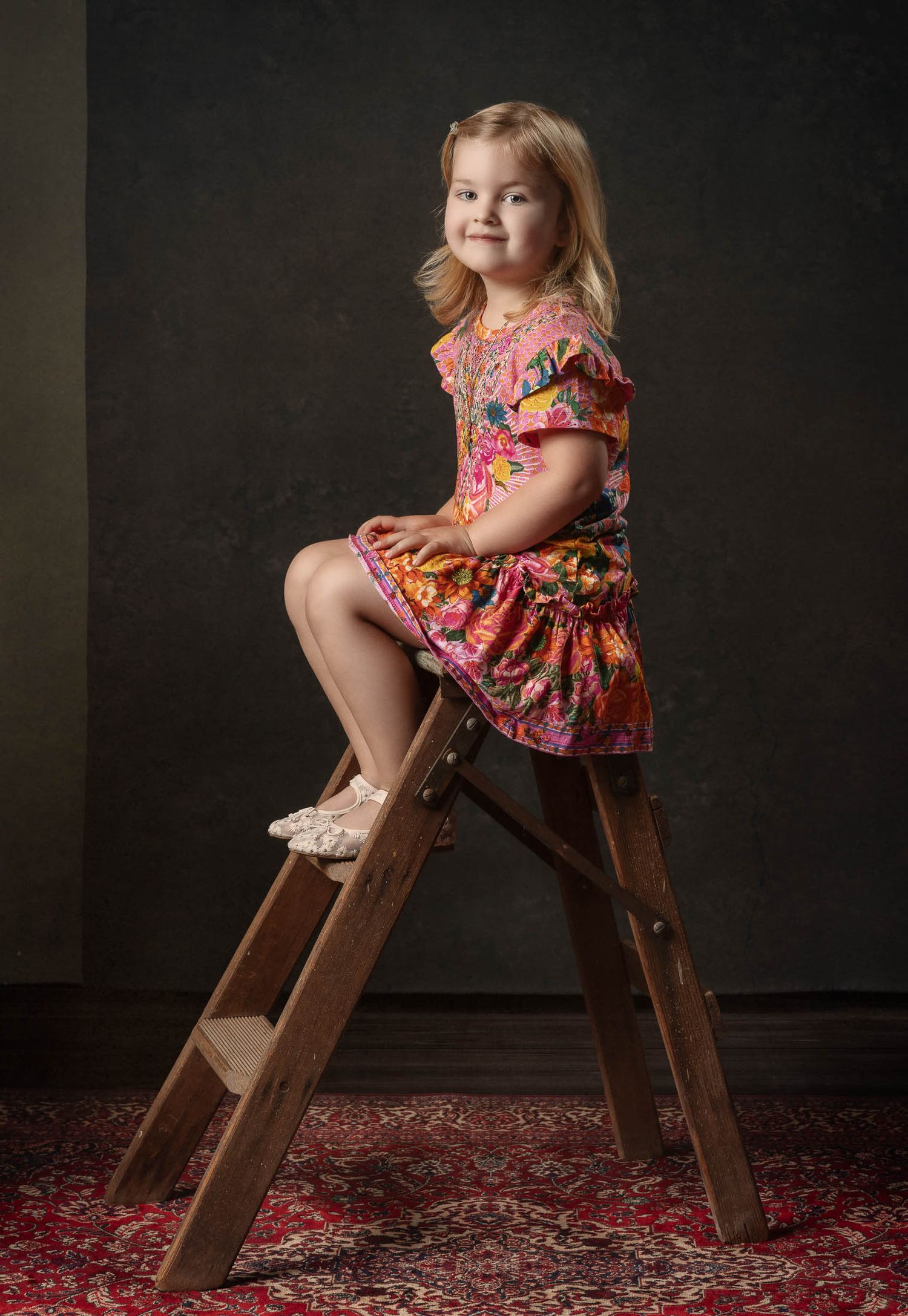 Smiling young girl in a colorful floral dress sits on a vintage wooden step ladder over a red patterned rug against a dark backdrop.
