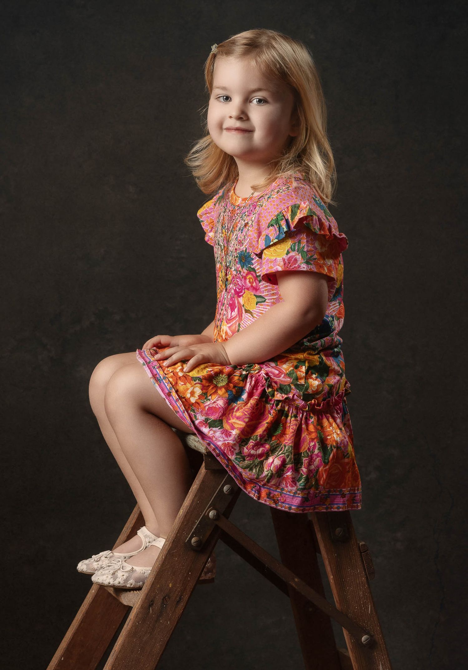 Painterly portrait smiling young girl in a colorful floral dress and sparkly sandals sits on a wooden ladder against a dark studio backdrop.