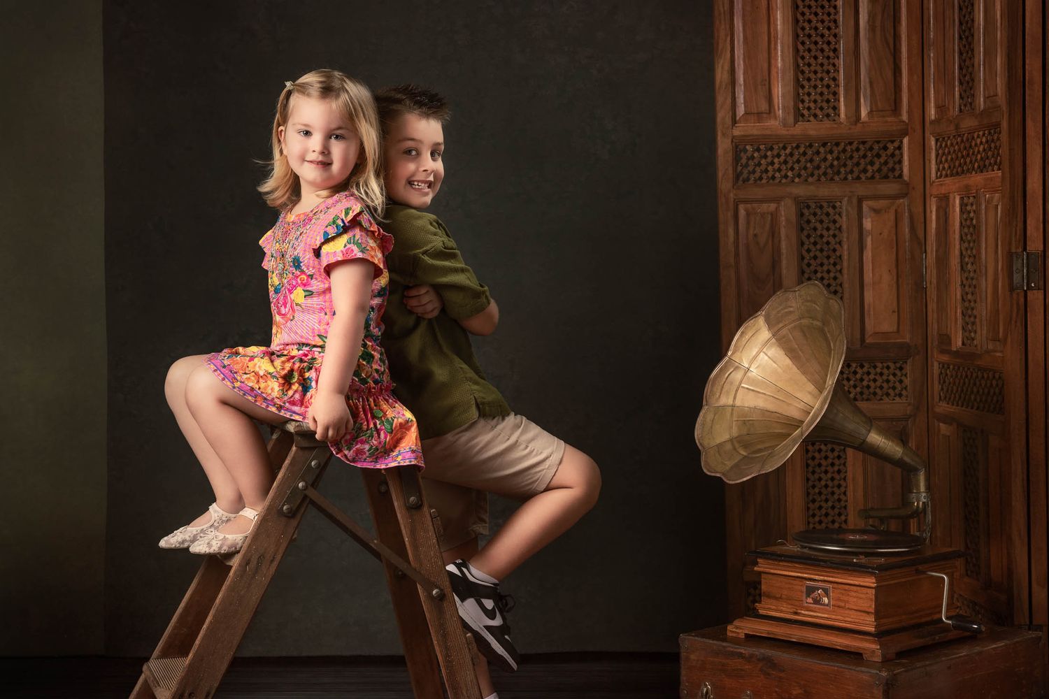 Smiling girl in a floral dress and boy in green shirt sit back to back on a wooden ladder beside a vintage gramophone.