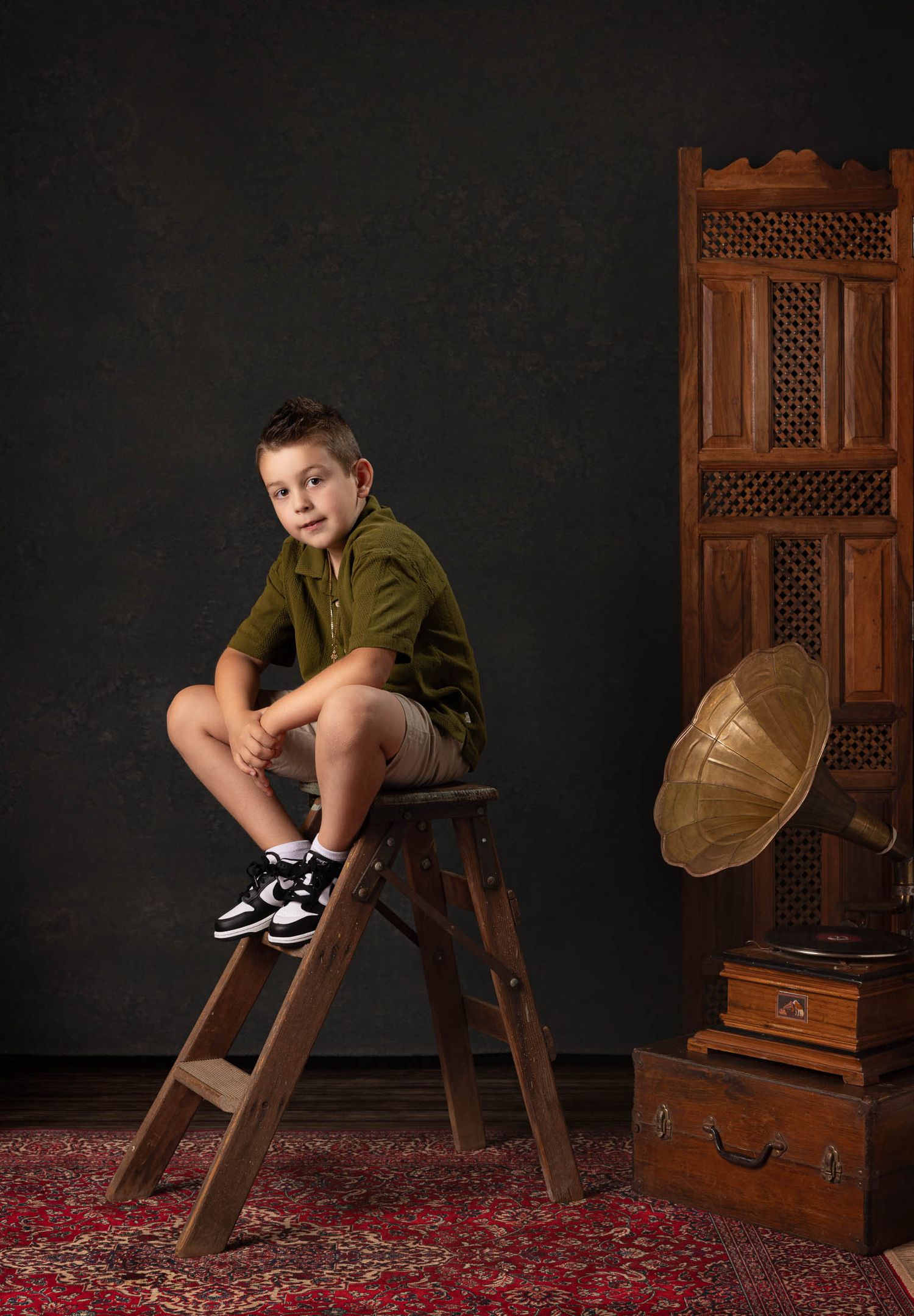 Young boy in an olive green shirt and shorts sits on a wooden stepladder beside an antique gramophone in a vintage studio portrait.