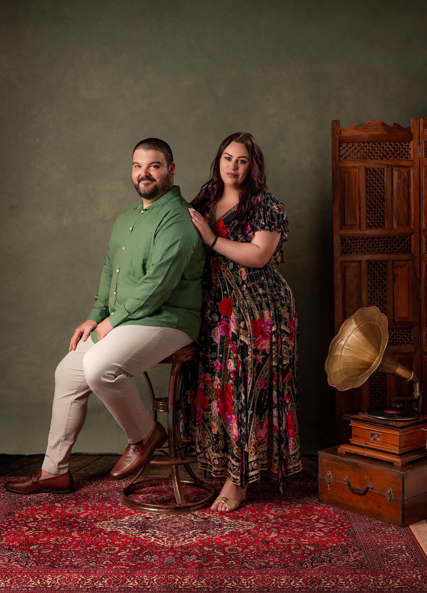 Bearded man in a green shirt sits on a stool beside a woman in a floral maxi dress, posing near a vintage gramophone on a red rug.