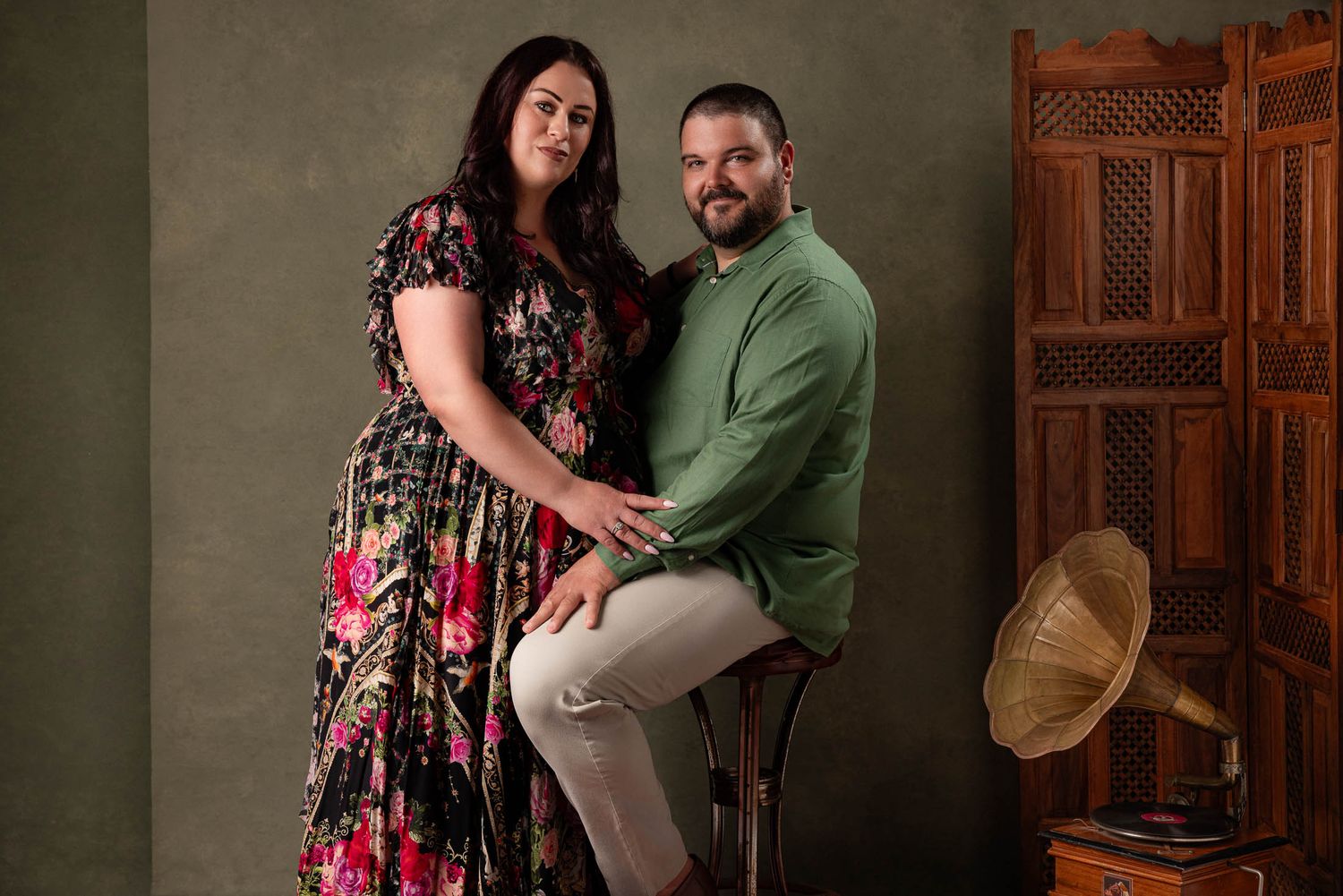 Smiling couple in a studio portrait, she in a floral maxi dress and he in a green shirt seated on a stool beside a vintage gramophone.