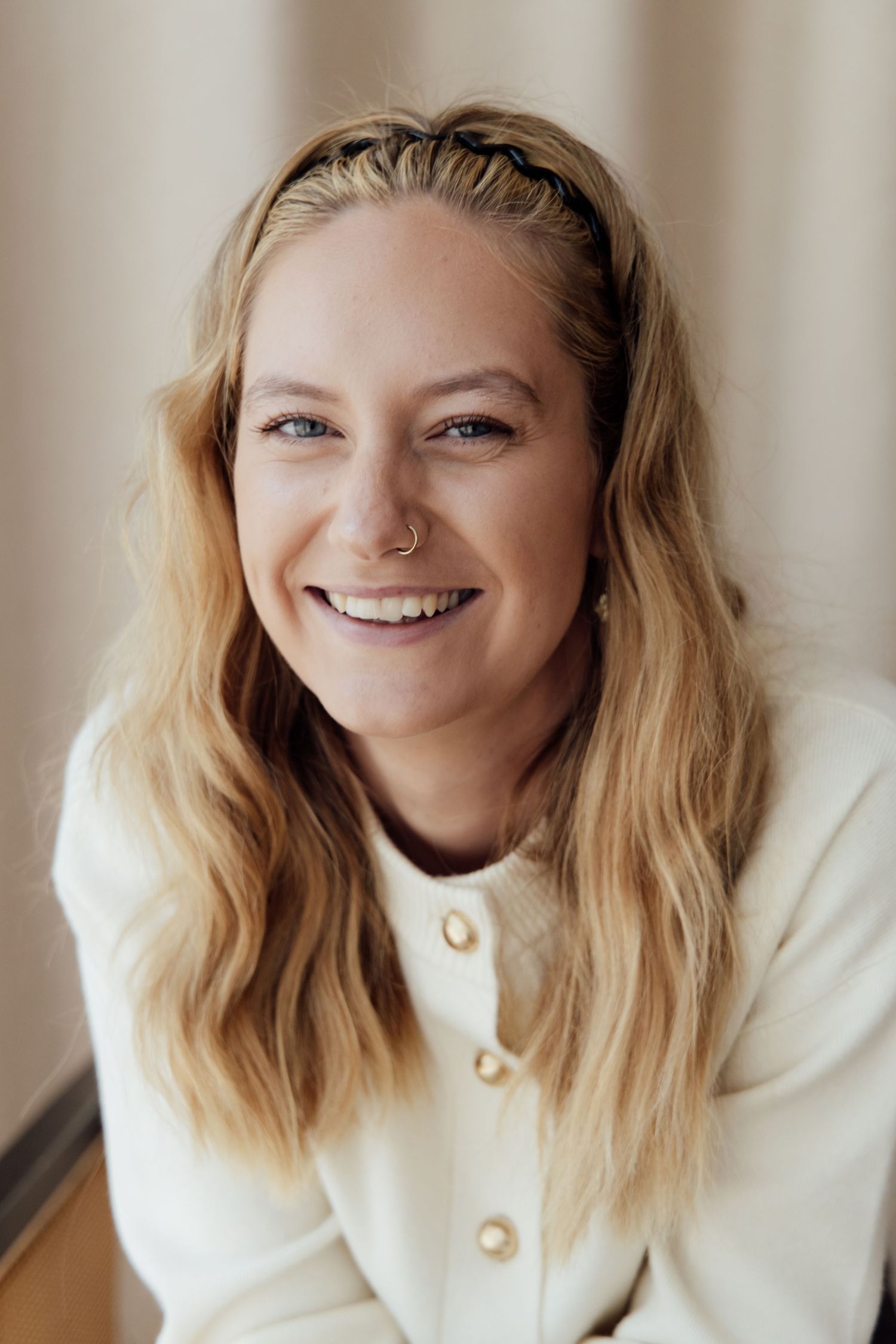 Smiling blonde woman with nose ring wearing white blazer and braided headband, portrait photo.
