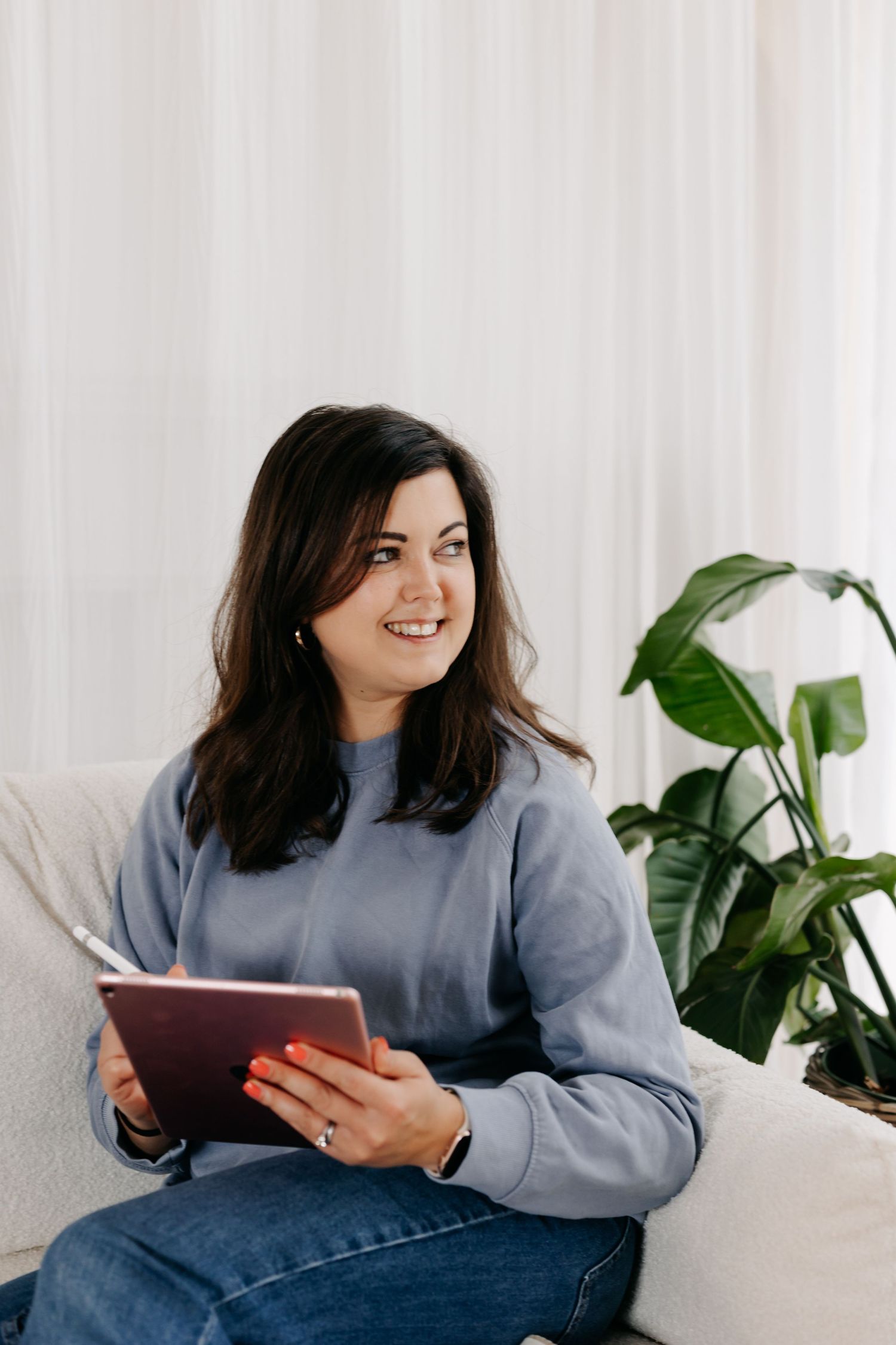 Smiling young woman sitting on a couch holding a tablet, wearing a gray sweatshirt and jeans in a bright living room.