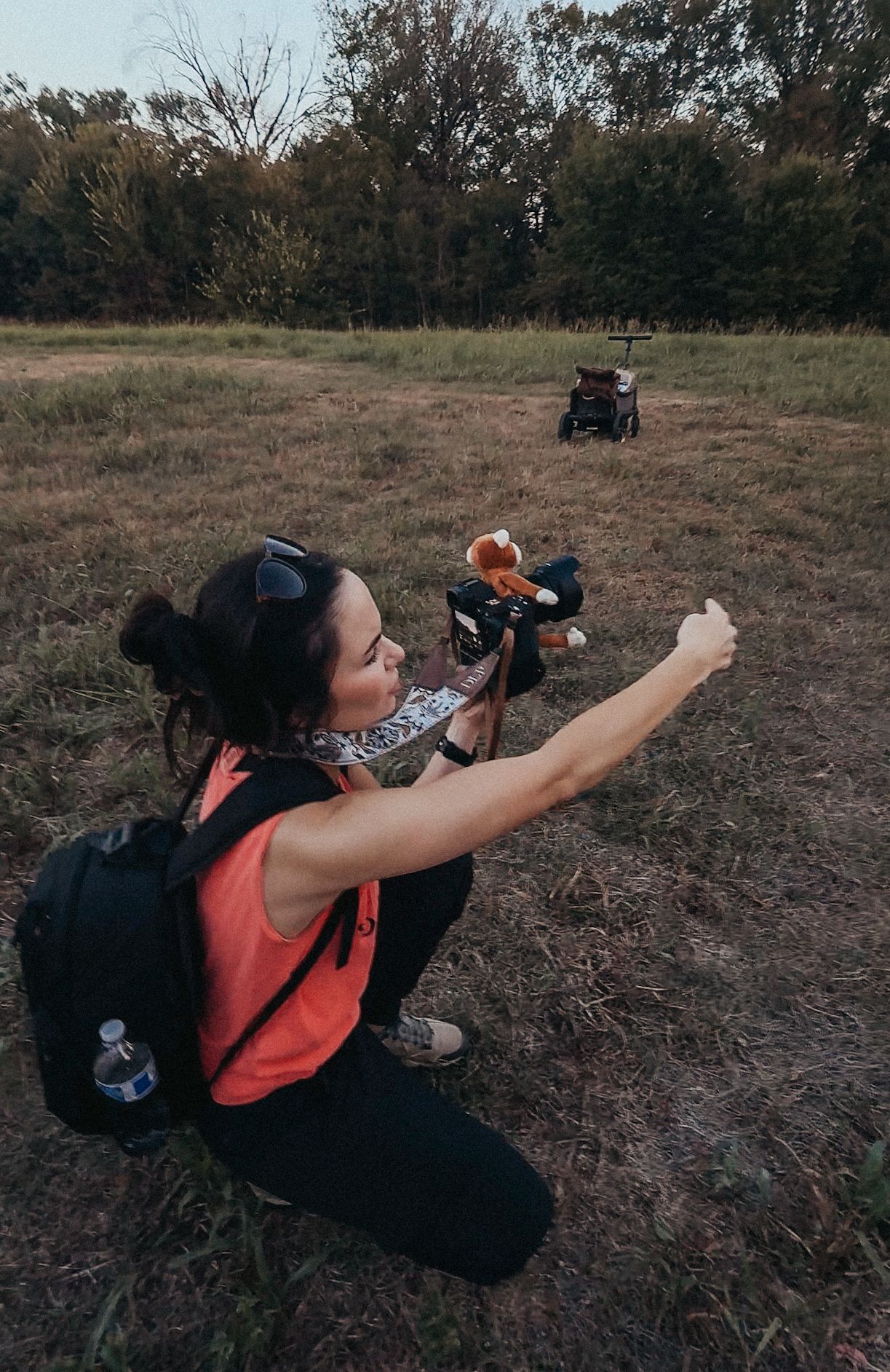 Woman with backpack kneeling in field taking selfie with drink, ATVs and trees visible in background.