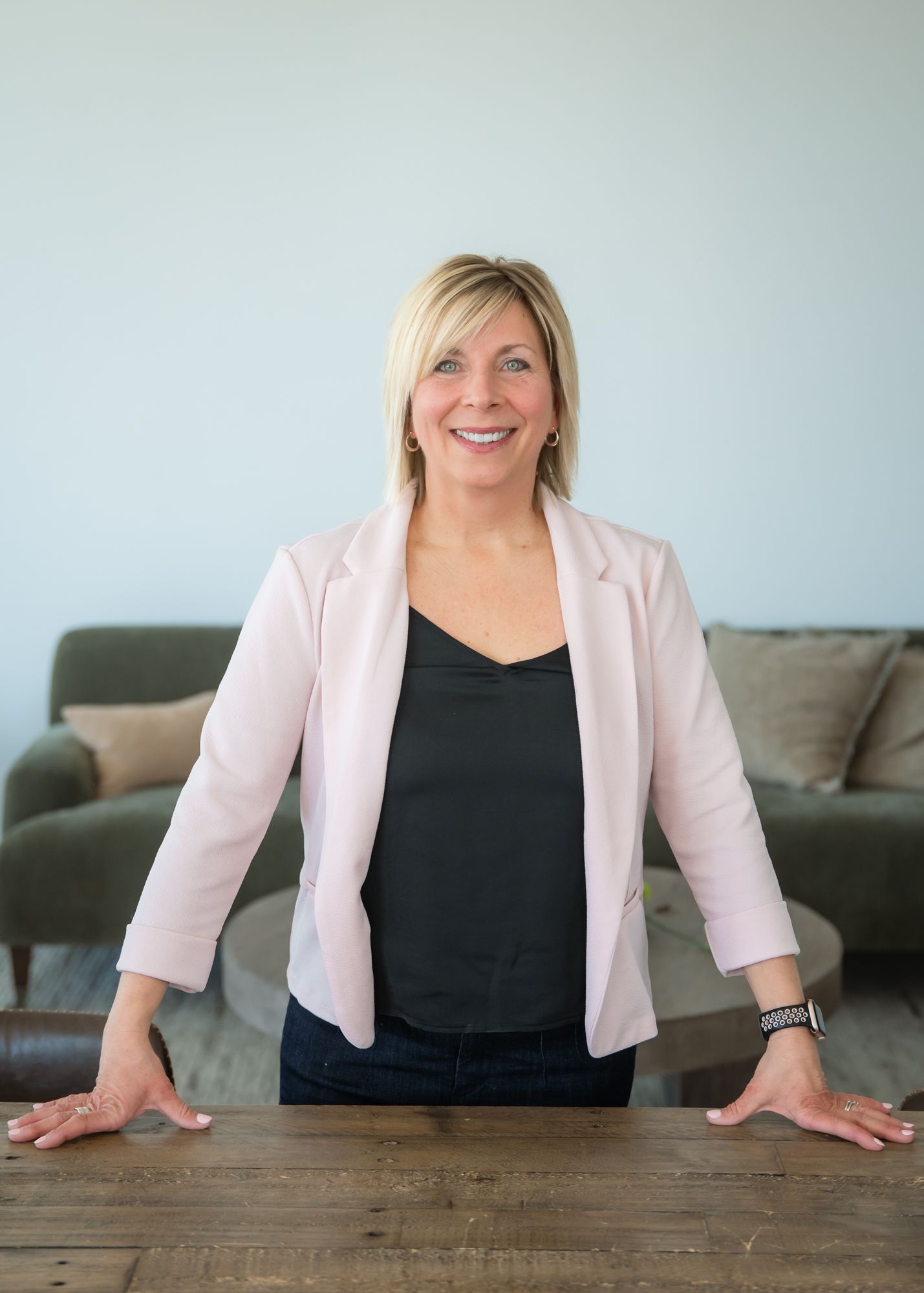 A smiling blonde woman in a pink blazer and black top stands confidently at a wooden table in a modern office.