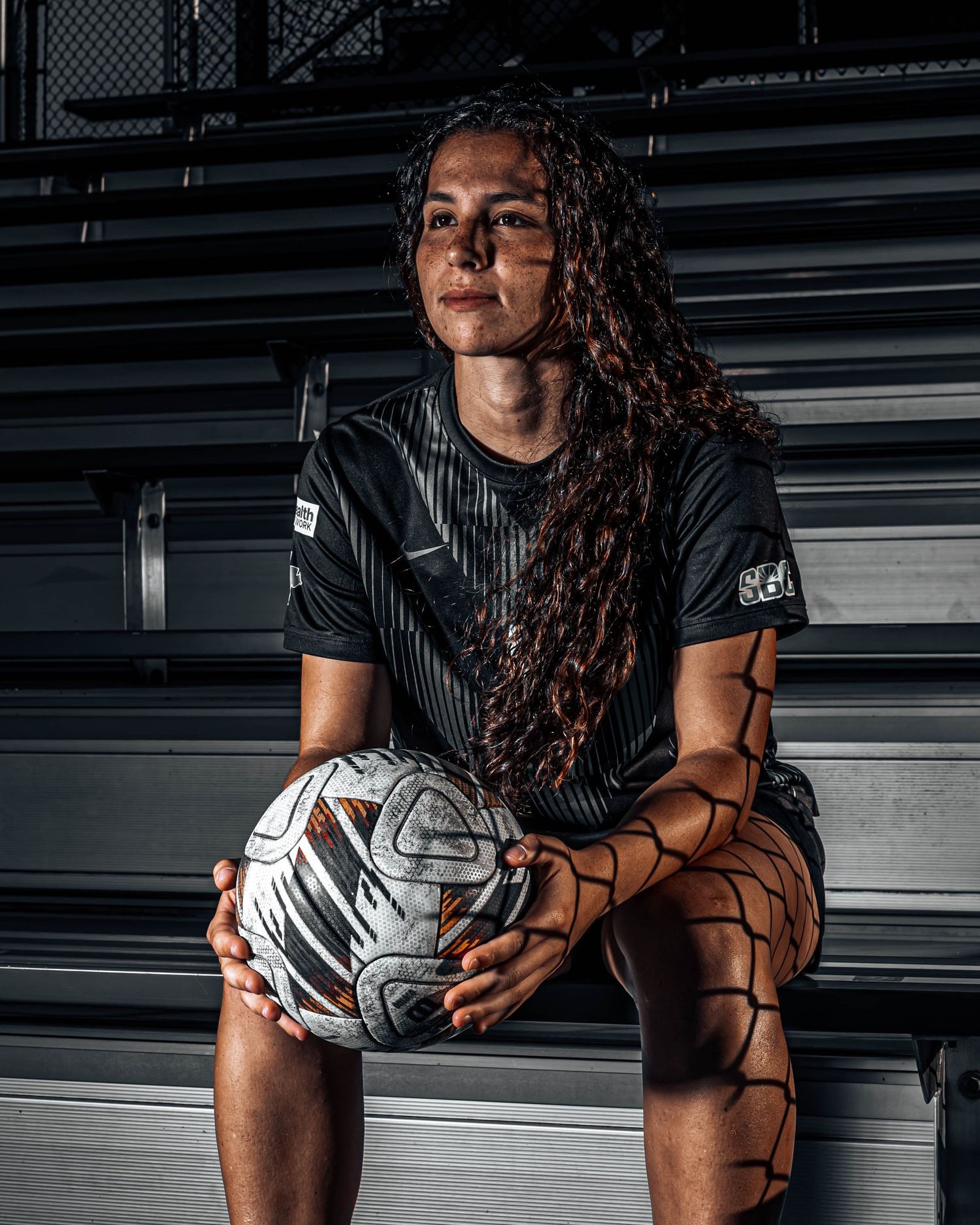Female soccer player in black jersey holds a ball while sitting on bleachers with dramatic lighting.