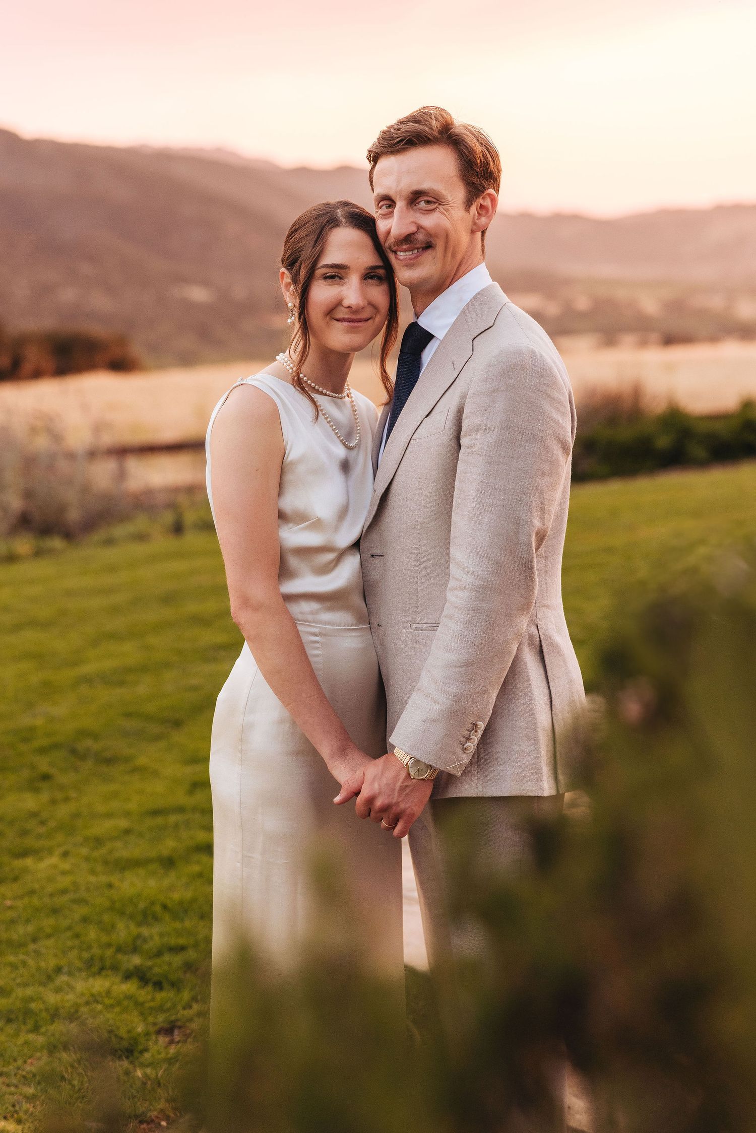 Couple embracing at golden hour near a wooden fence in a rural setting, bride in white dress, groom in tan suit.
