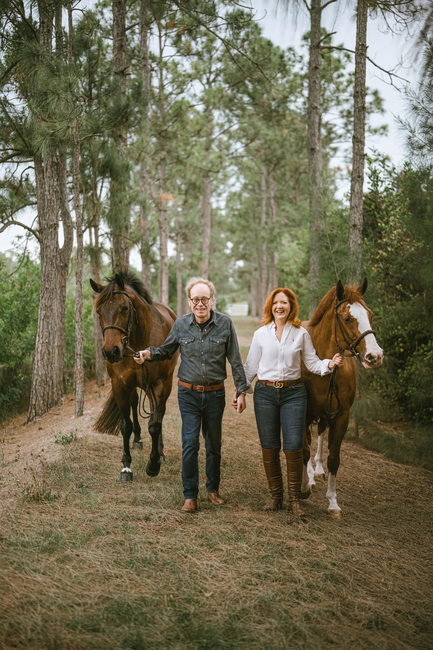 Couple walking with two brown horses along a tree-lined dirt path in a serene outdoor setting.