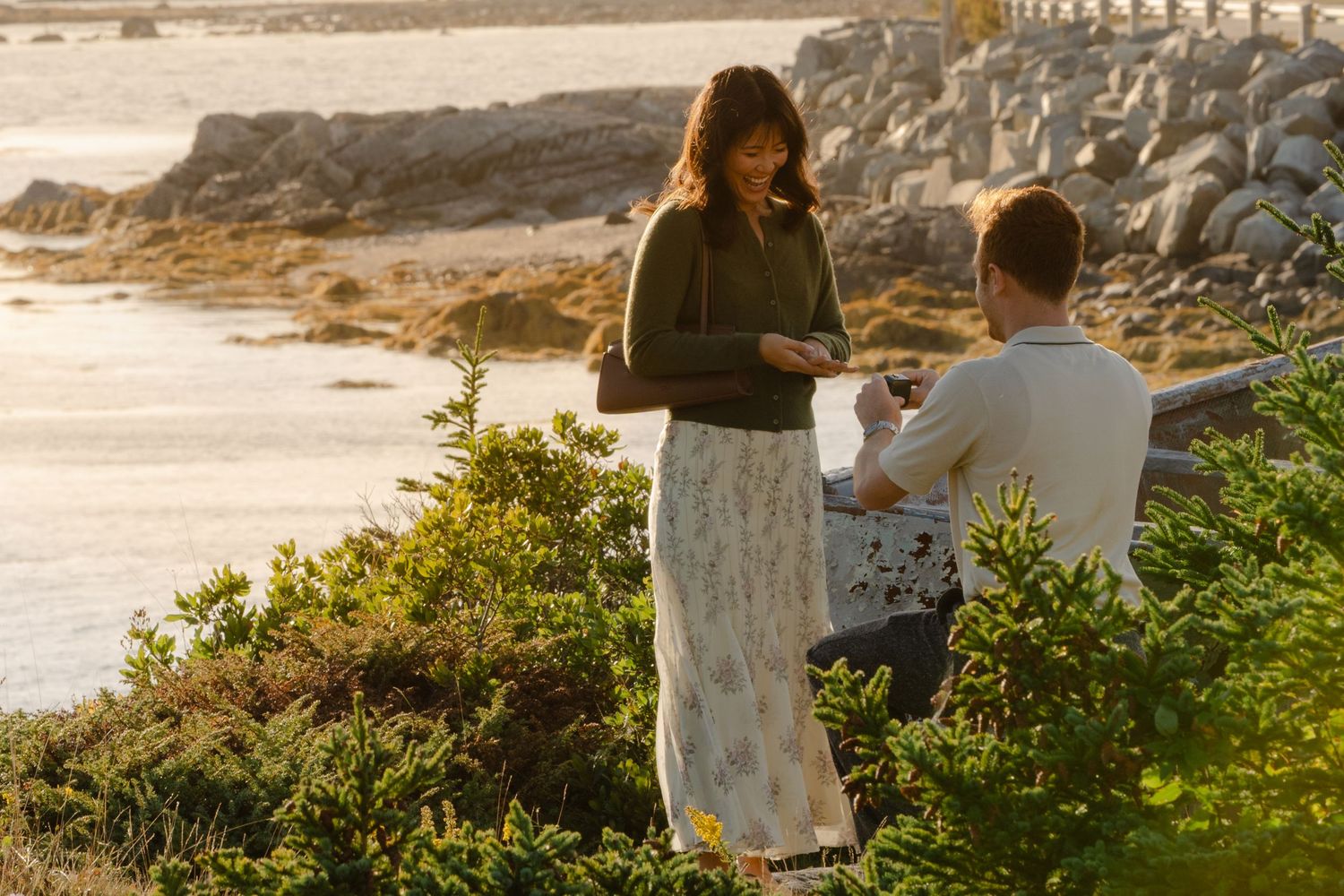 Man proposes to woman near rocky shoreline at sunset, surrounded by lush greenery and ocean views.