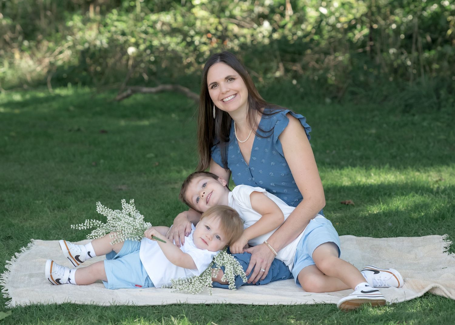 A smiling woman sits on a blanket outdoors with two young boys, surrounded by green grass and trees.