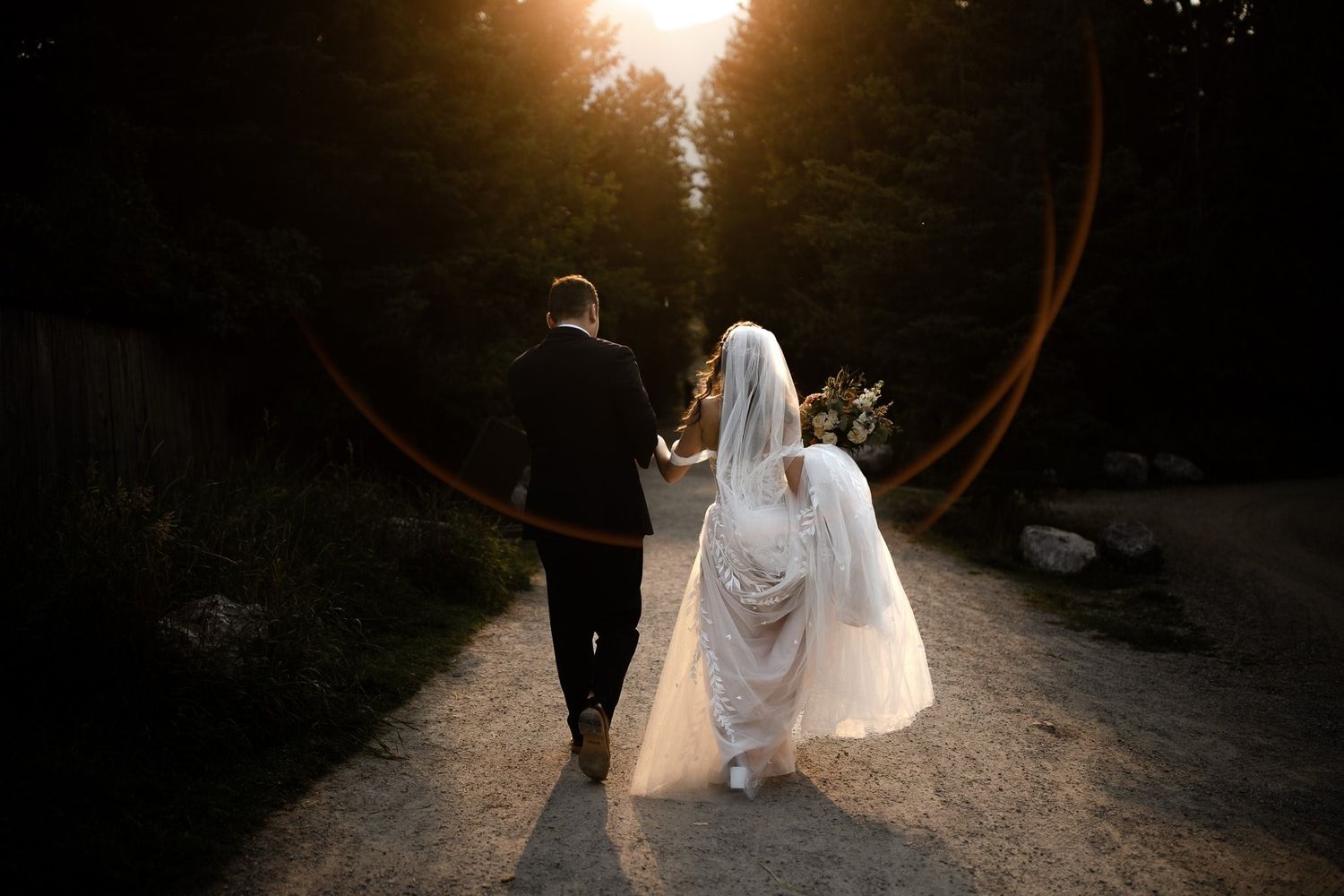 Bride and groom walking hand in hand down a sunlit forest path, by a Calgary wedding photographer