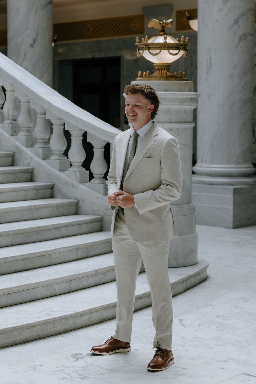 Smiling groom in a beige suit stands confidently on marble stairs in an elegant venue.