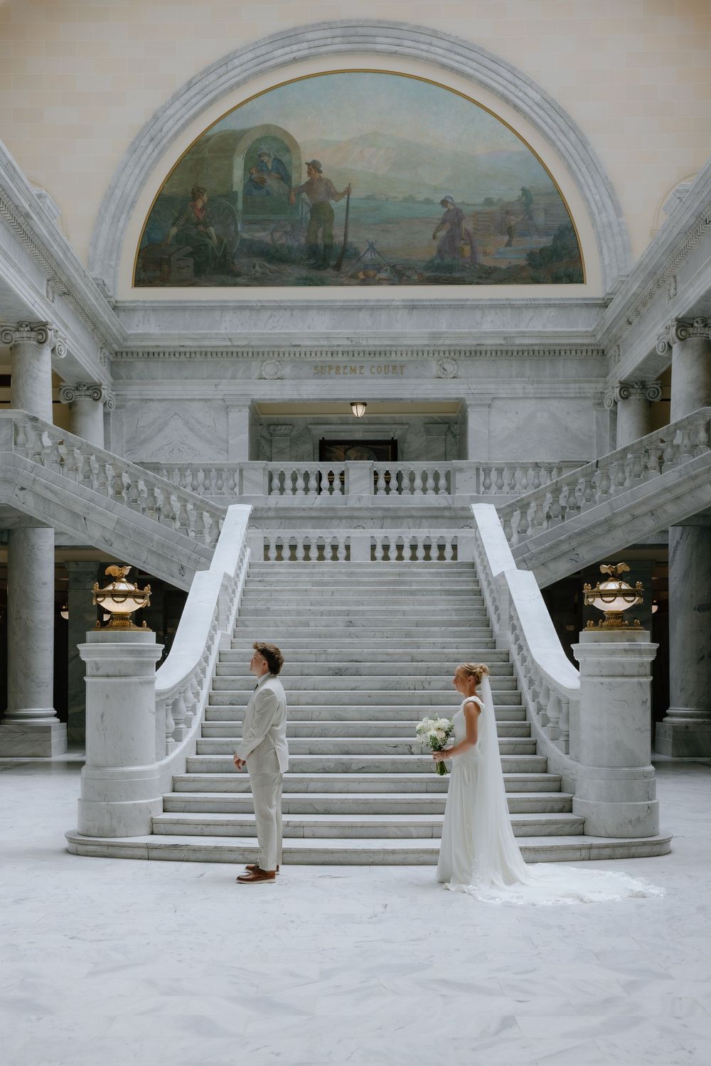 Bride and groom standing before grand marble staircase beneath a classical mural in capitol building.