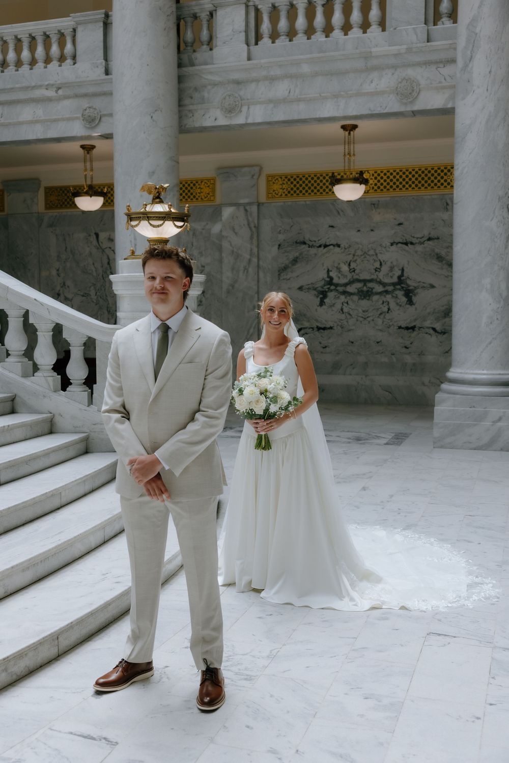 Groom stands in foreground while bride with bouquet poses behind him on marble stairs of a grand building.