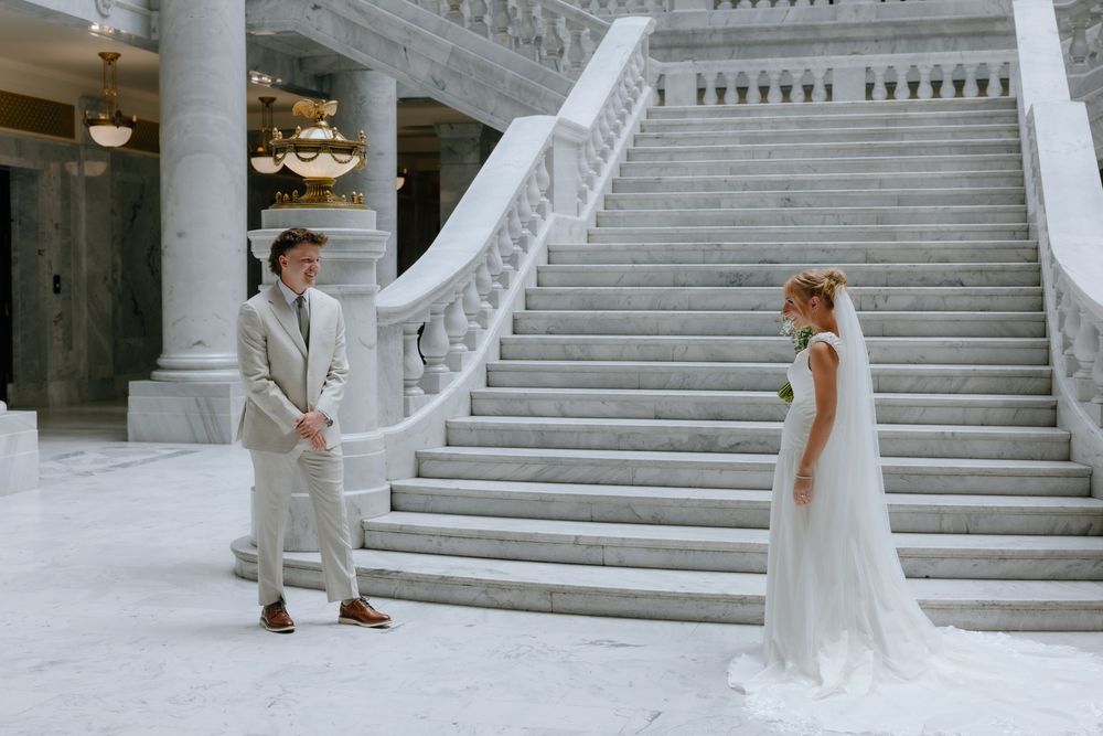 Groom in beige suit sees bride in white gown for the first time at marble staircase.
