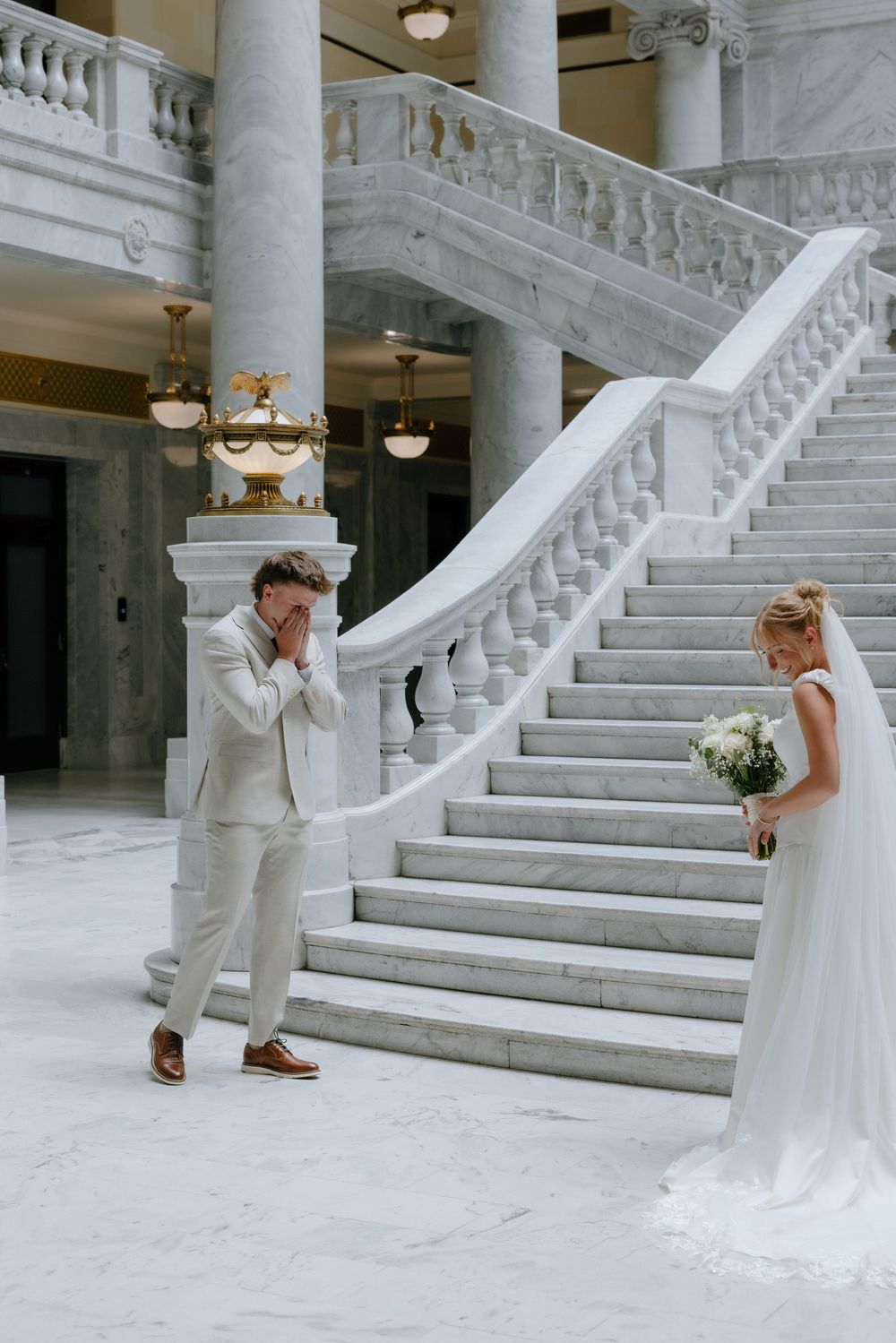 Emotional groom covers his face seeing bride in white gown at marble staircase first look.