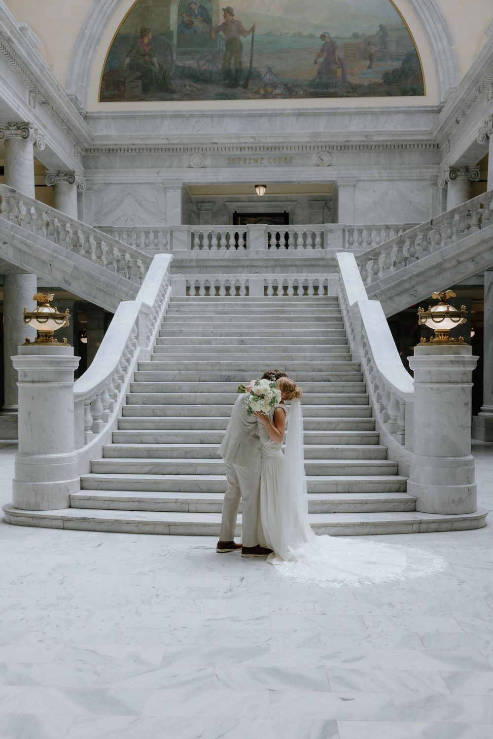 Bride and groom embrace at the base of a grand marble staircase with a mural above.