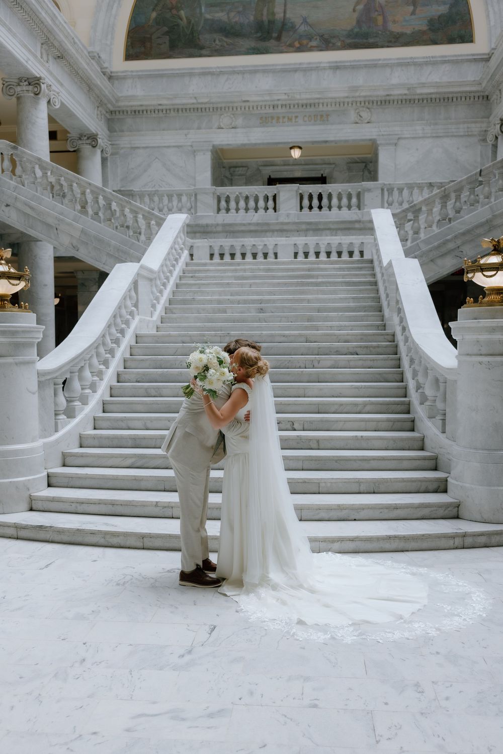 Bride and groom share a kiss at the base of grand marble staircase with white floral bouquet.