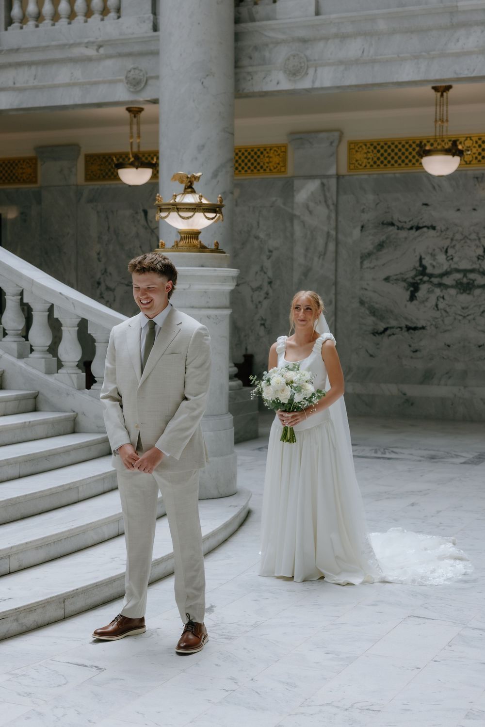 Groom smiles at first look as bride approaches with white bouquet on marble staircase at venue.