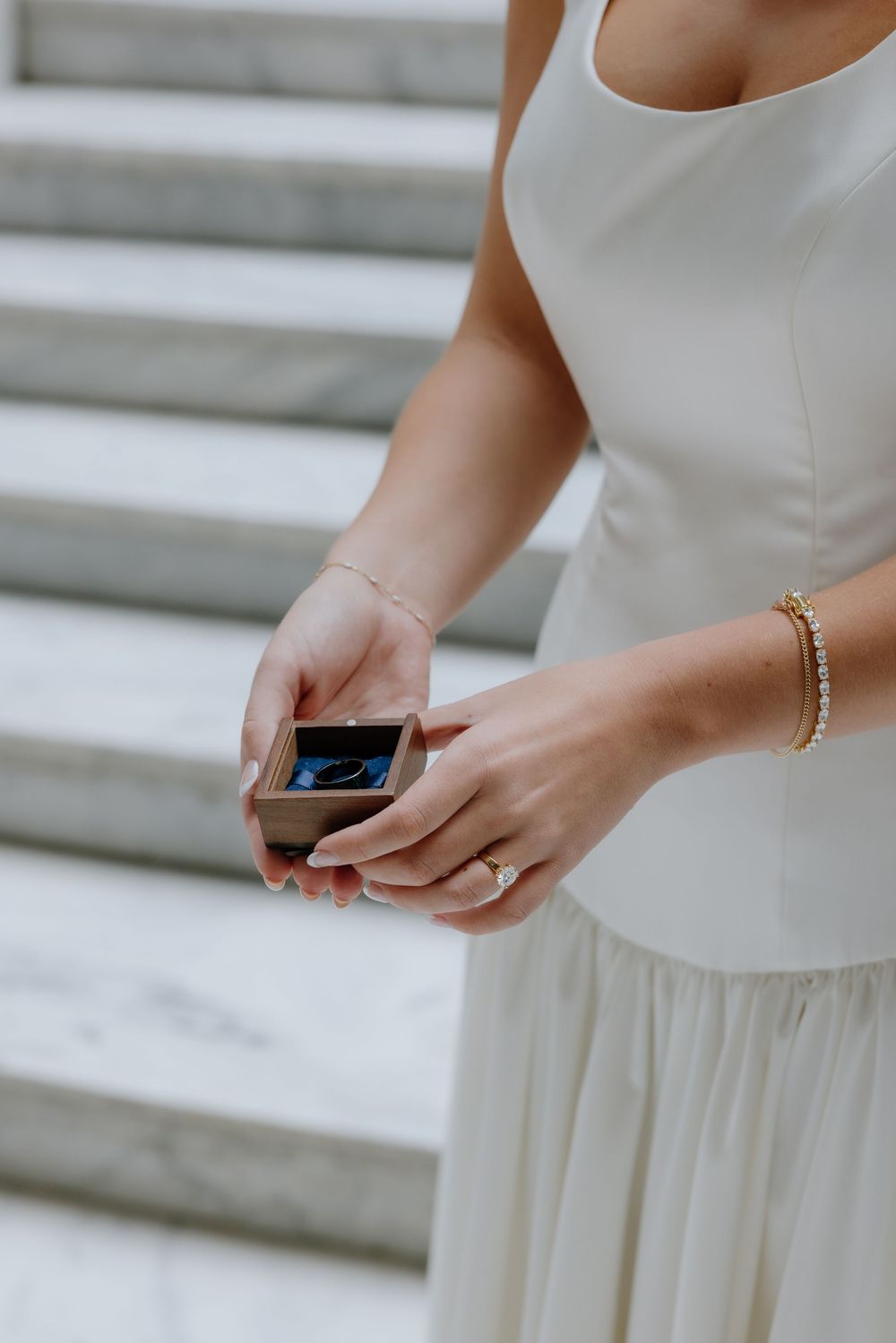 Bride in white gown holds wooden ring box on marble steps wearing gold bracelet and engagement ring.