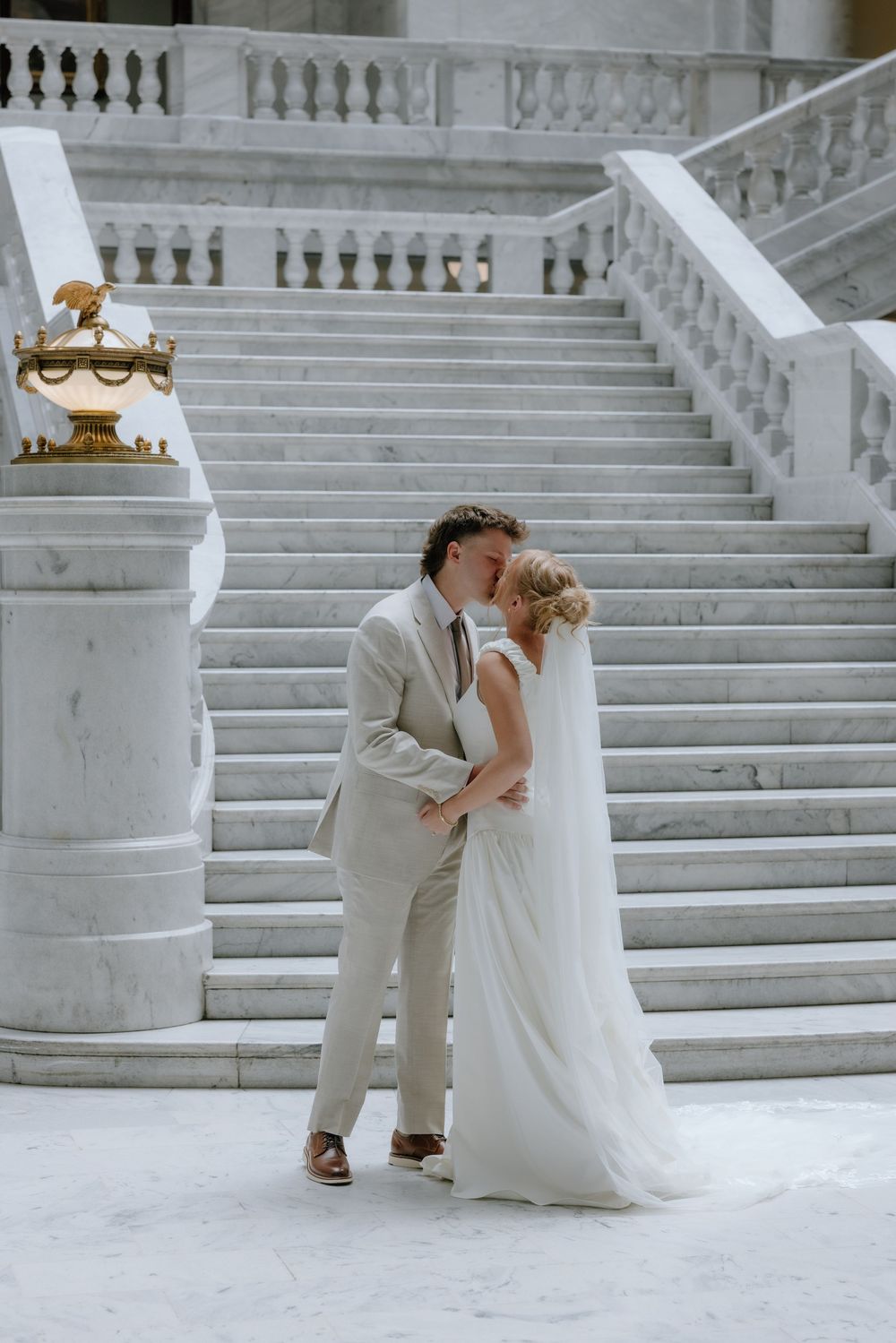 Bride and groom share romantic kiss at base of grand marble staircase.