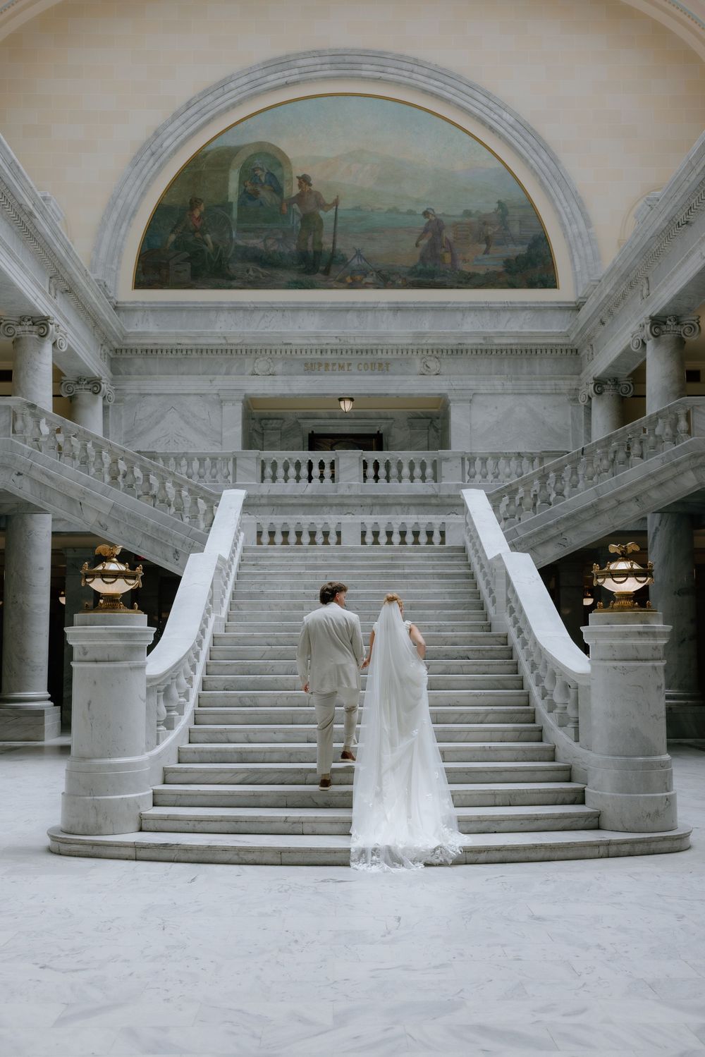 Bride and groom walk hand-in-hand toward grand marble staircase beneath stunning rotunda mural in capitol building.