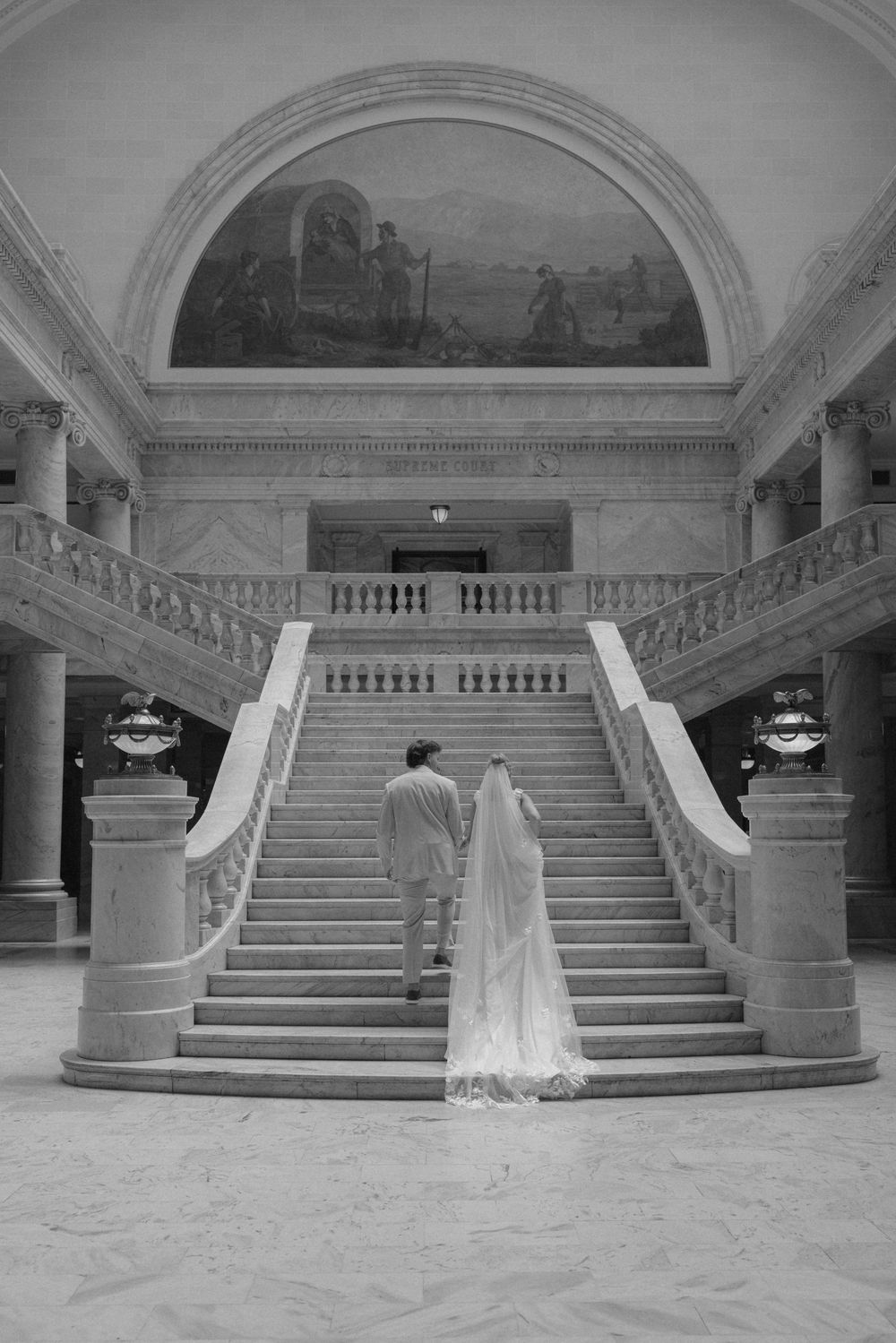 Bride and groom walk toward grand marble staircase beneath ornate mural in black and white wedding photo.