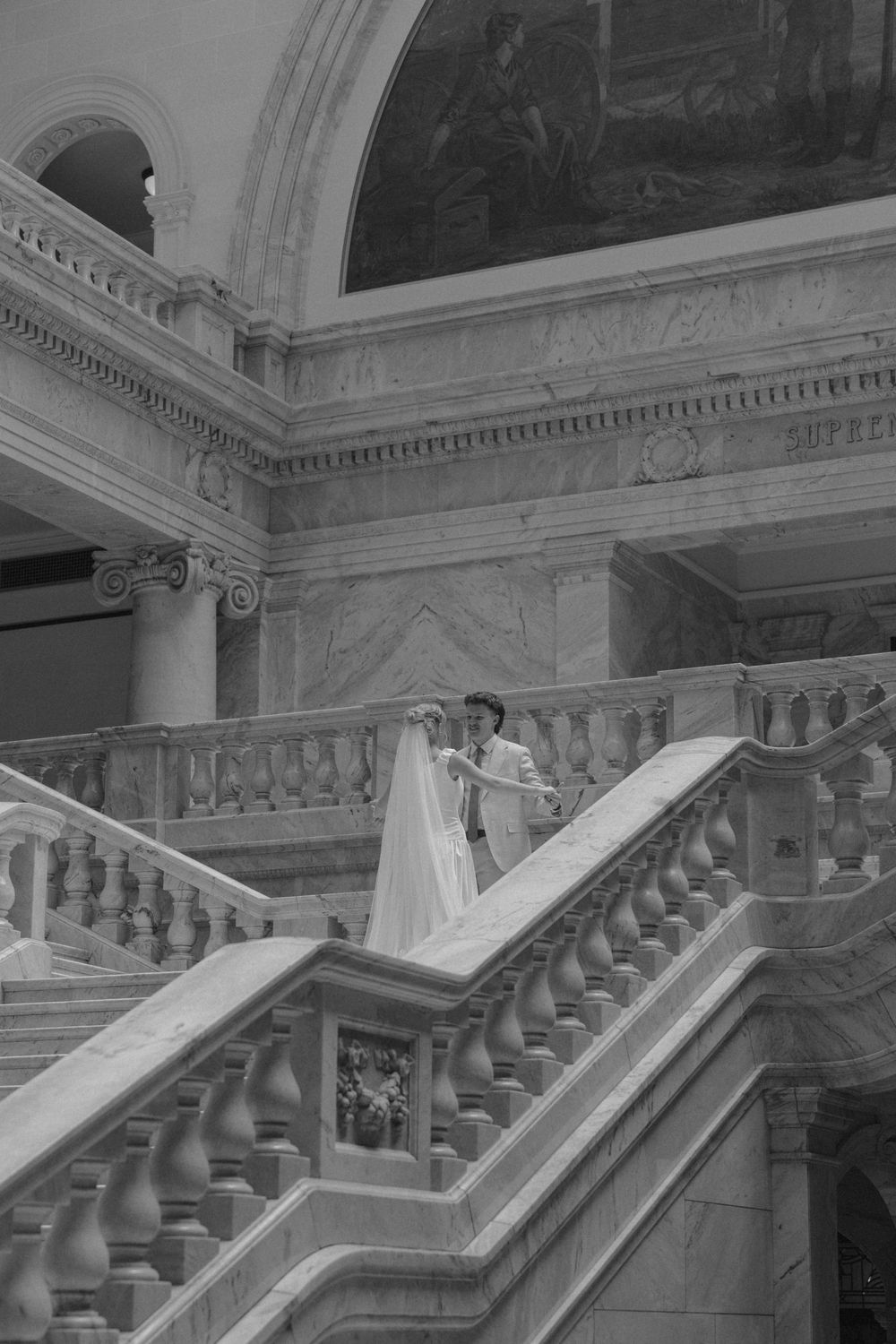 Black and white photo of bride and groom on grand marble staircase with arched murals in stately building.