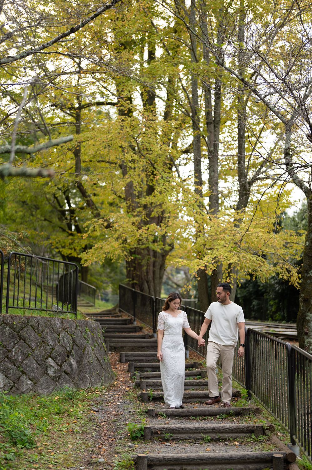 Couple holding hands walk down wooden steps along a tree-lined path surrounded by yellow autumn foliage and a stone wall.