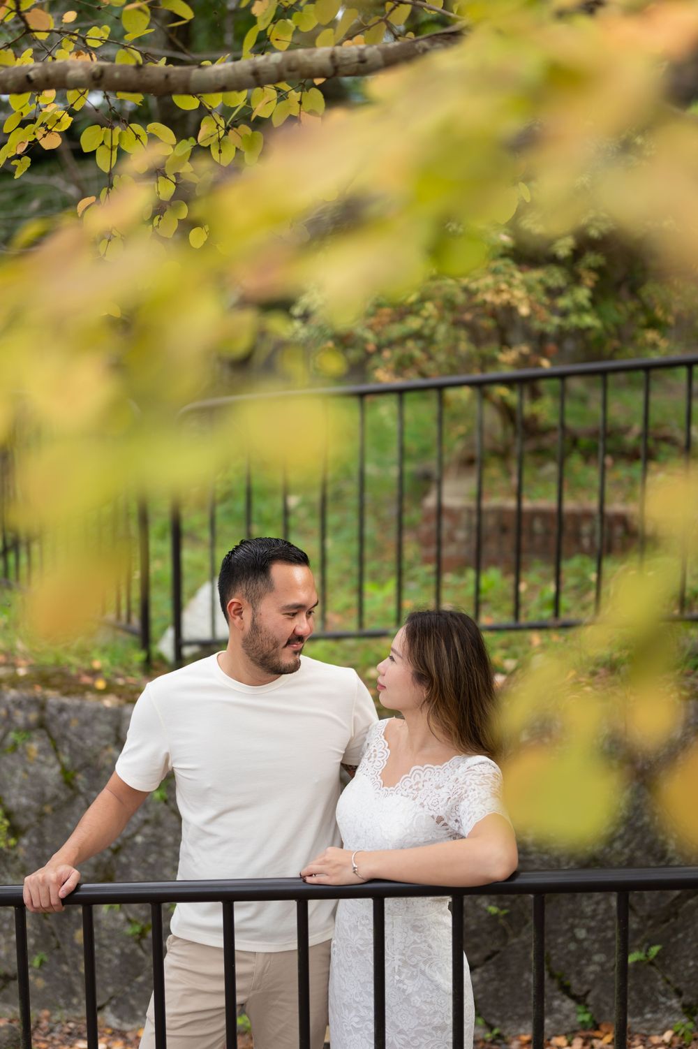 Smiling couple in casual white outfits share a tender moment beside a black metal railing, framed by blurred golden autumn leaves.
