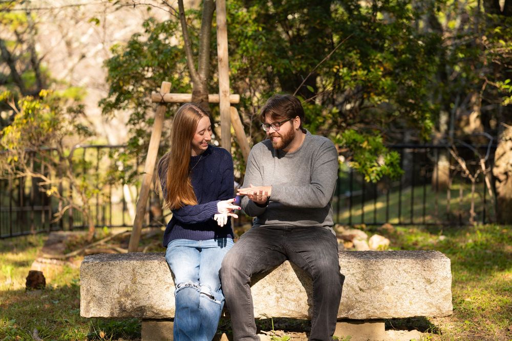 A young woman and a bearded man in glasses sit on a stone bench in a sunlit park, smiling as they look at something in their hands.