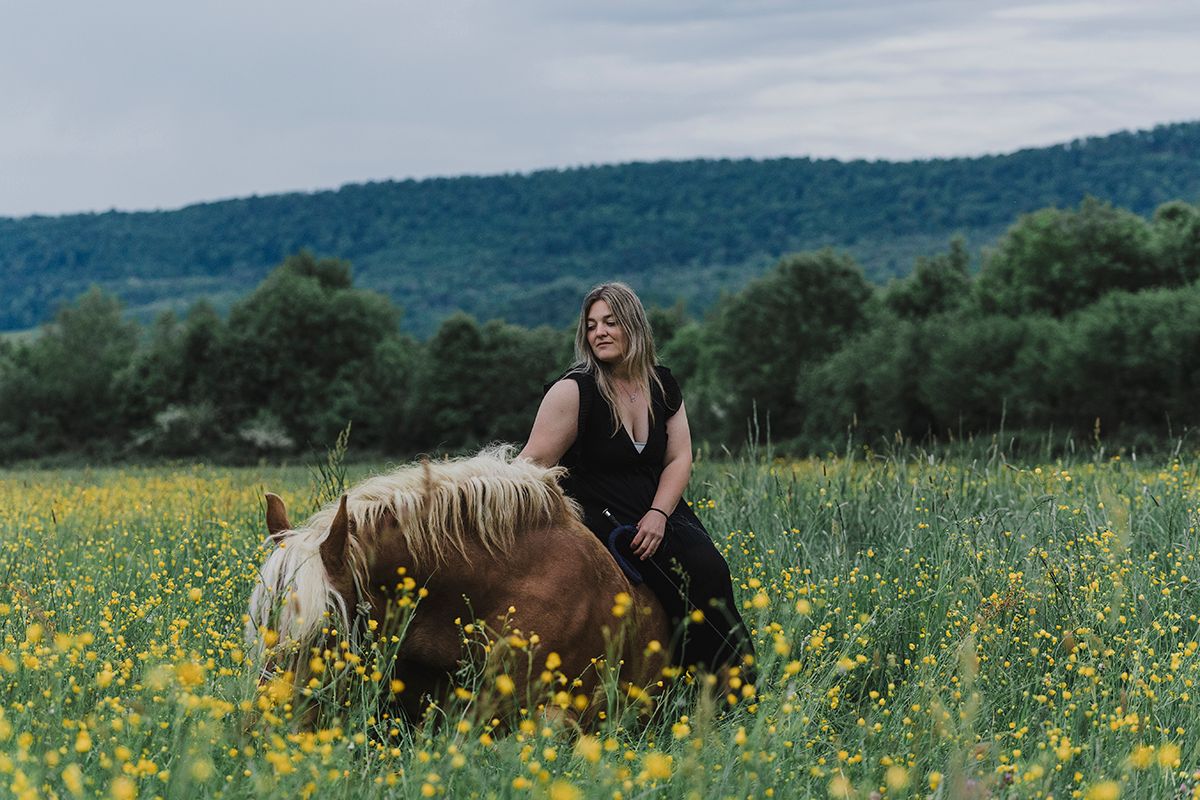 Woman in black dress riding a chestnut horse through a field of yellow wildflowers with green hills behind.