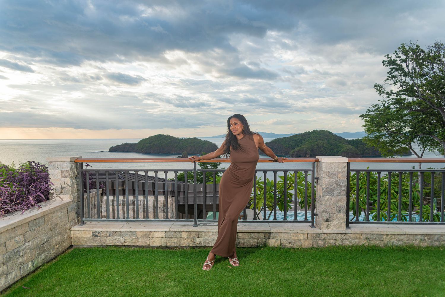 Woman in a brown dress poses at a stone terrace railing with a sweeping ocean and green hills view at sunset.