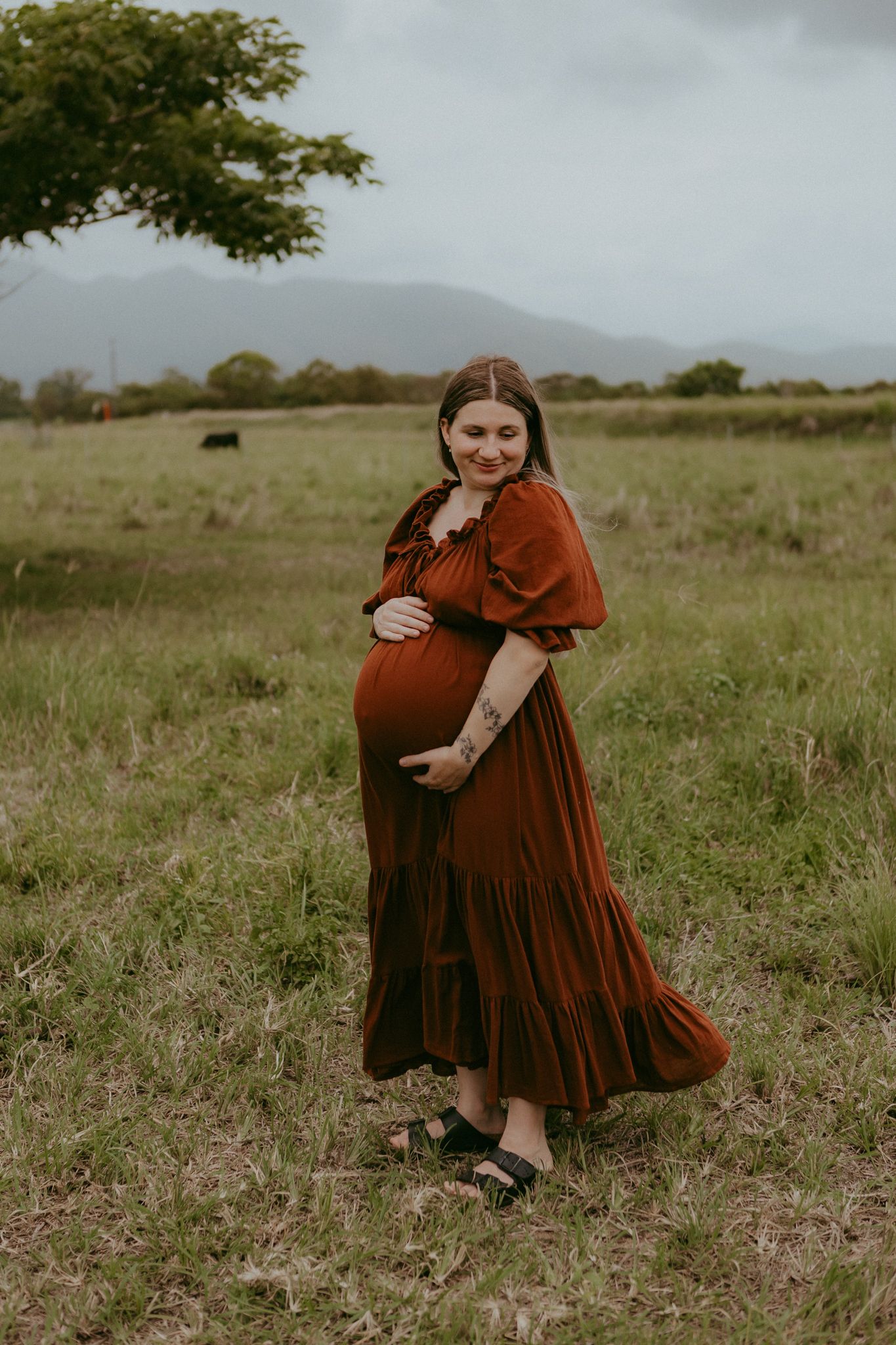 A pregnant woman in a rust-colored dress cradles her baby bump in a scenic open green pasture.