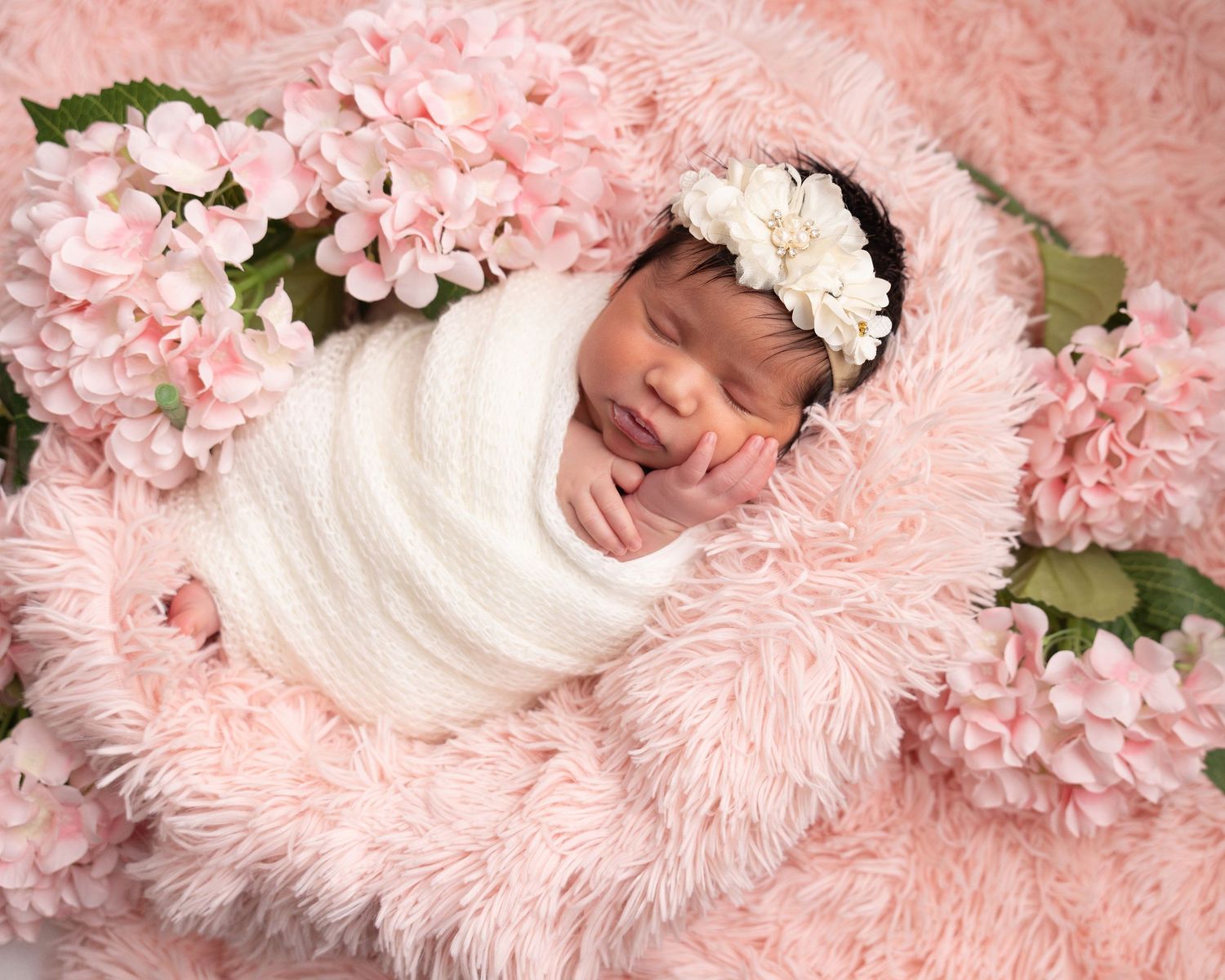 Newborn baby wrapped in white swaddle with floral headband sleeping on pink fur surrounded by pink flowers.